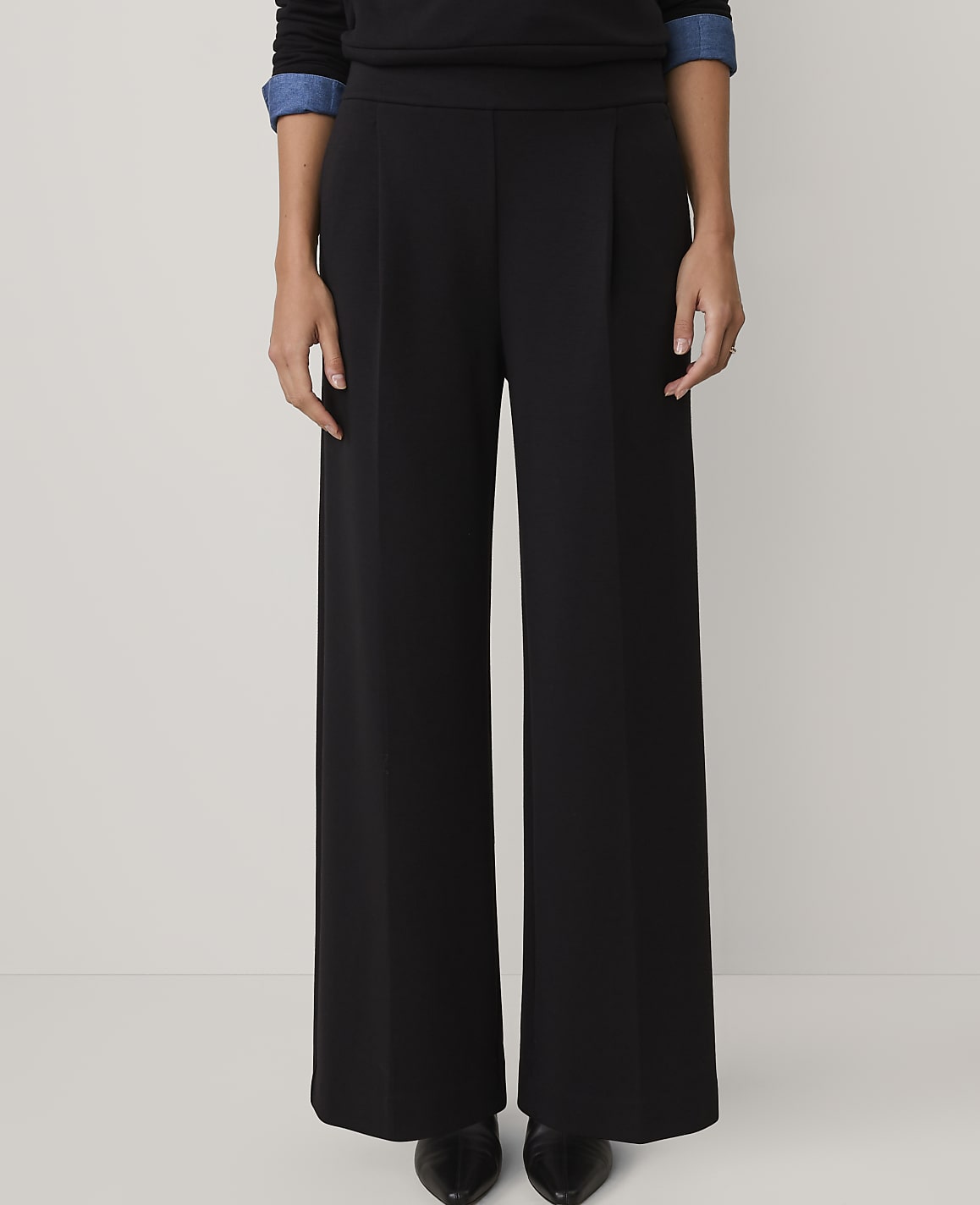 A person wearing black wide-leg trousers and a dark top stands against a plain, light-colored background.