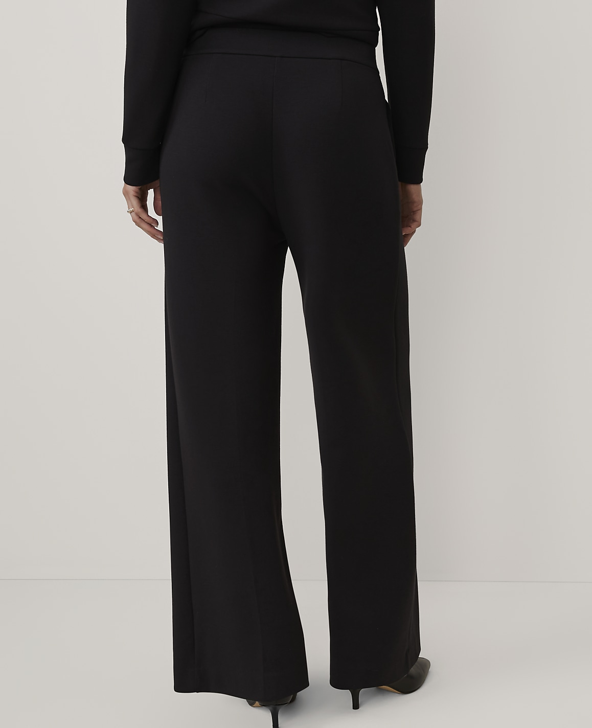 A person wearing a black long-sleeved top and wide-leg black trousers stands against a plain, light-colored background.
