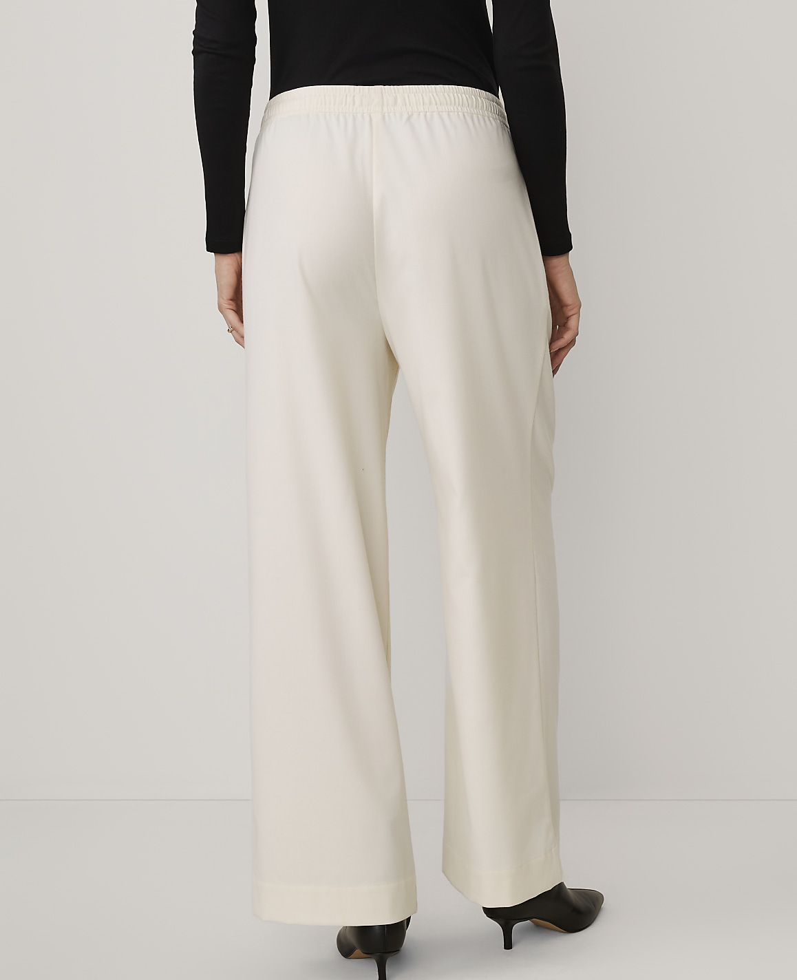 A person wearing a black long-sleeved top and wide-leg cream trousers stands against a plain light gray background.