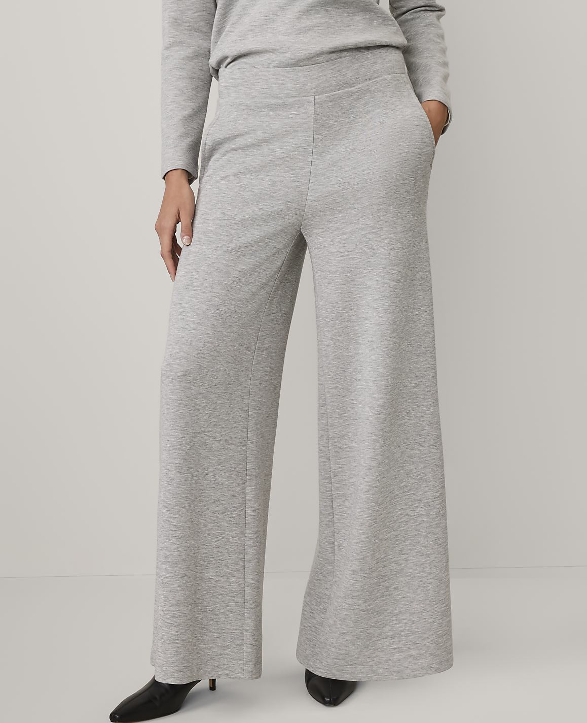 A person wears a light grey long-sleeved top and matching wide-leg trousers with black pointed heels against a plain, light-colored background.