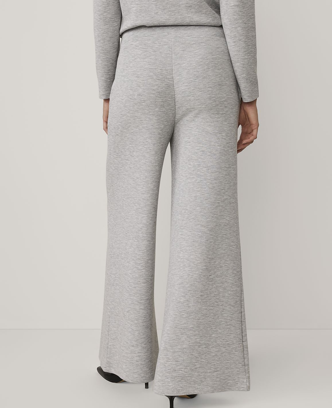 A person stands with their back to the camera wearing grey wide-leg trousers and a matching top against a plain white background.