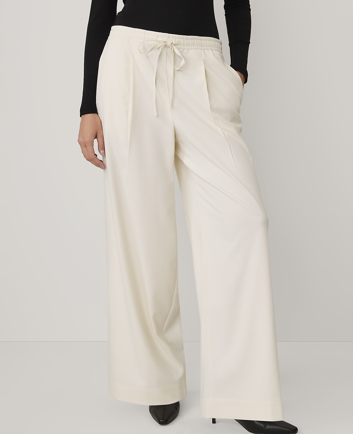 A person wearing a black top and wide-leg cream trousers stands against a plain white background.