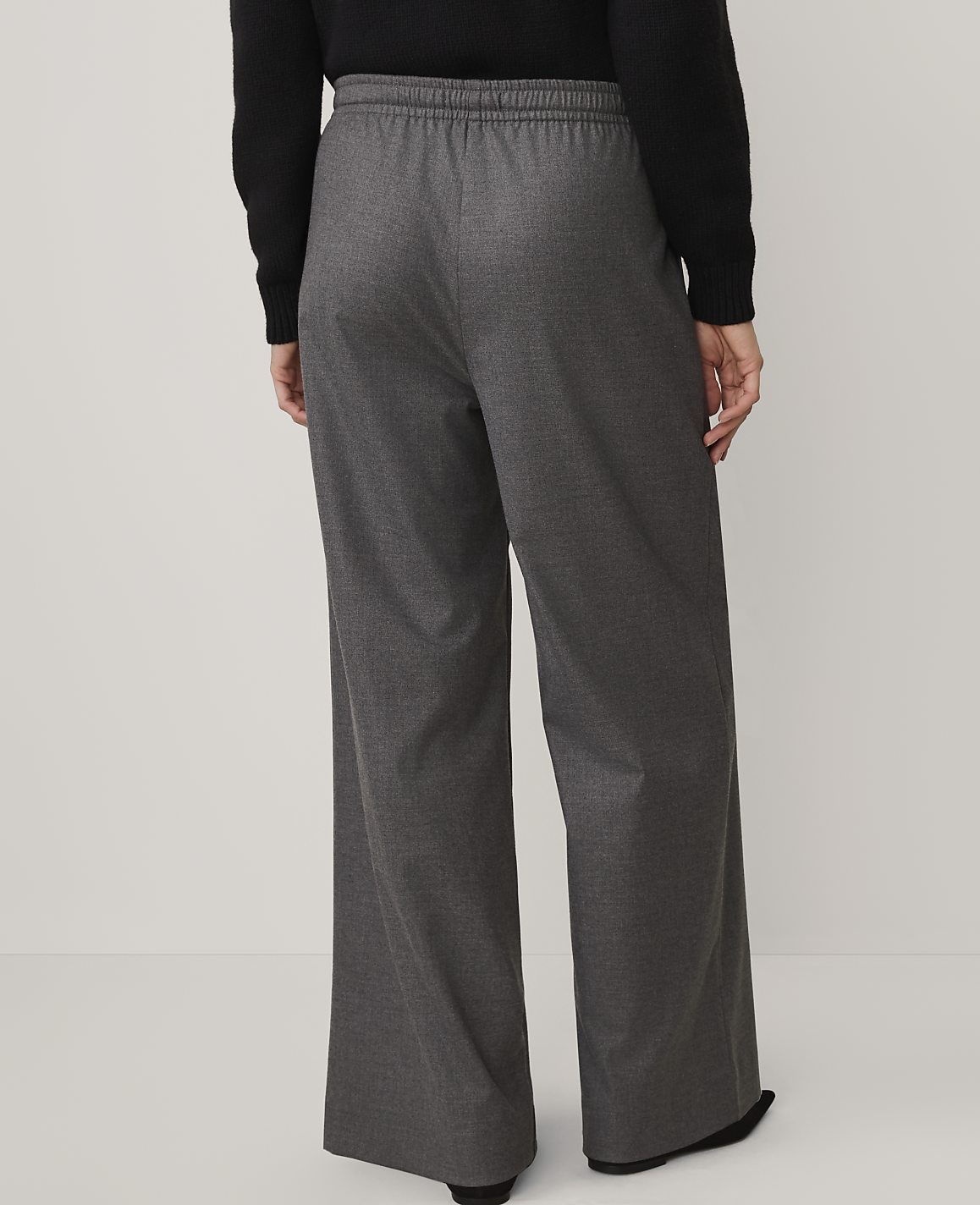 A person wearing a black top and wide-leg grey trousers stands against a plain, light-colored background.