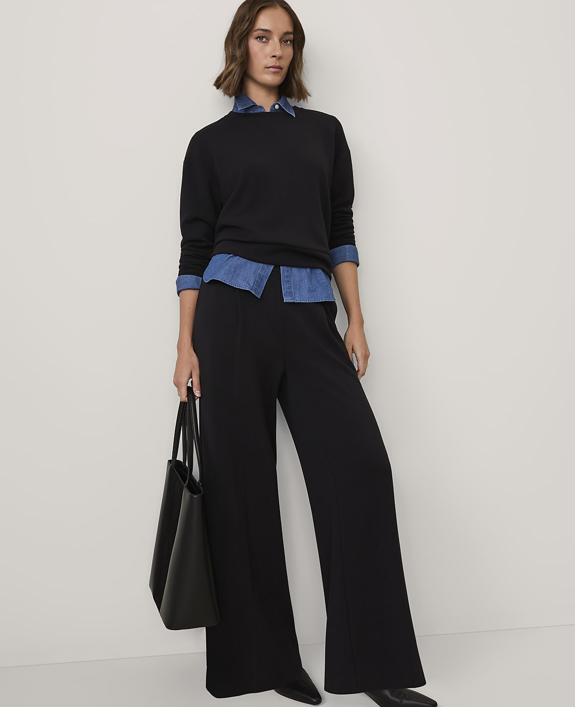 A woman wearing a black sweater over a blue denim shirt and wide-leg black trousers stands against a plain white background.