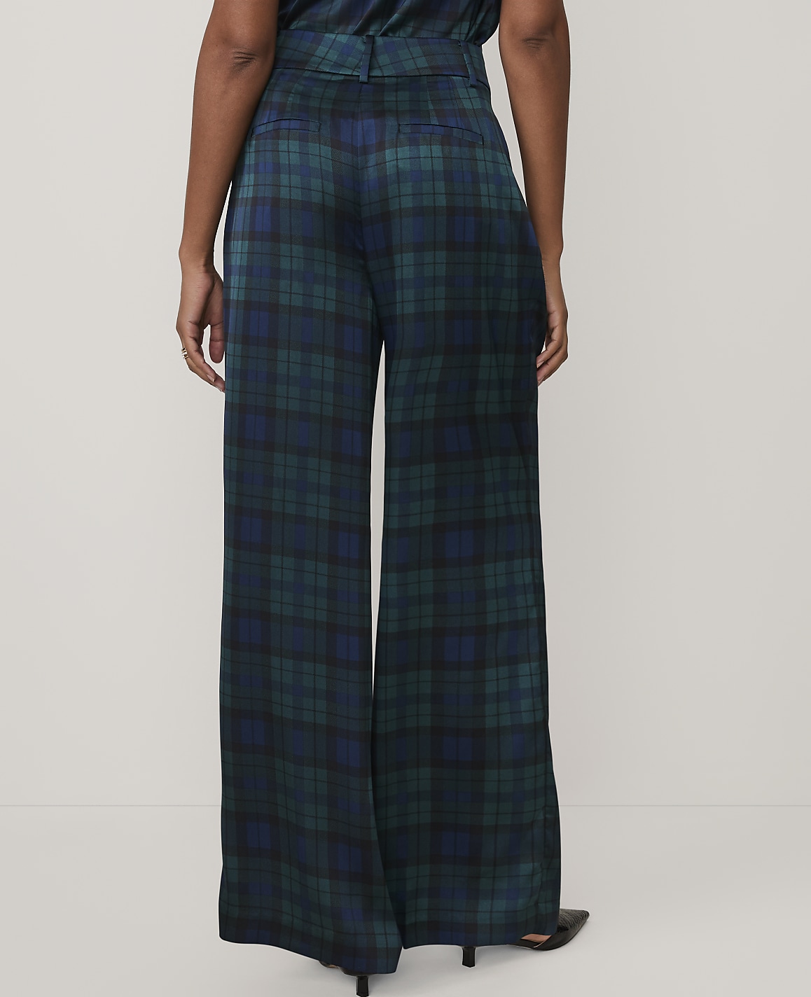 A person stands facing away from the camera wearing wide-leg, dark green and blue plaid trousers against a plain white background.