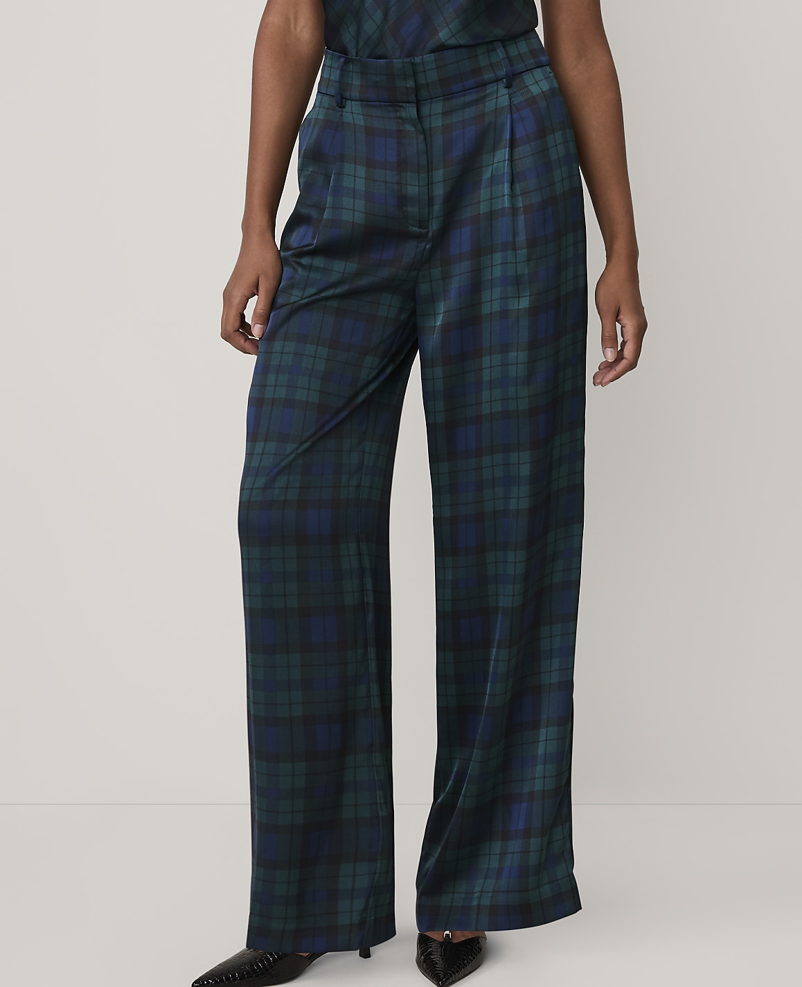 A person wears wide-leg, dark green and blue plaid trousers and black pointed shoes against a plain, light-colored background.