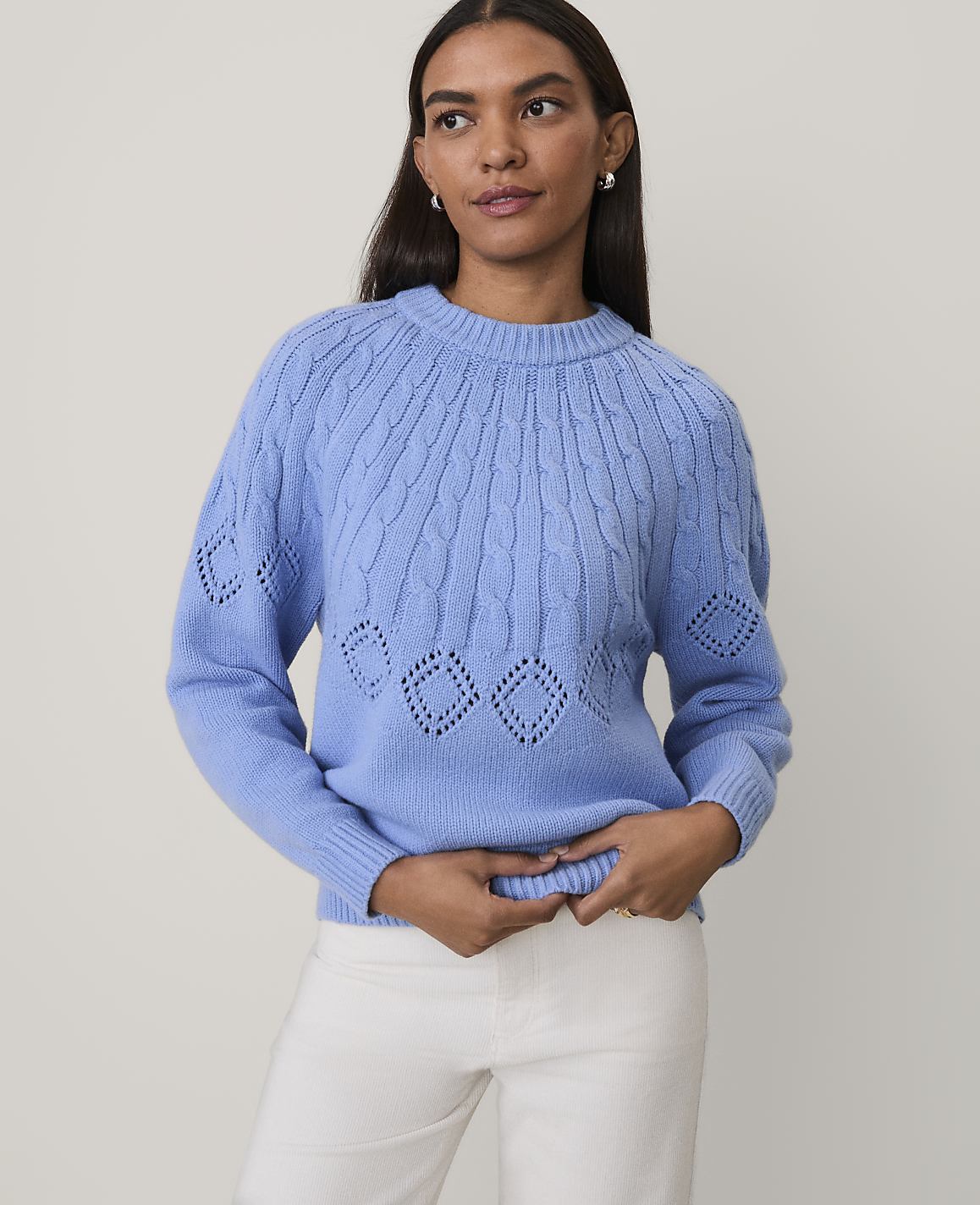A woman wearing a blue cable-knit sweater and white pants stands against a plain, light-colored background.