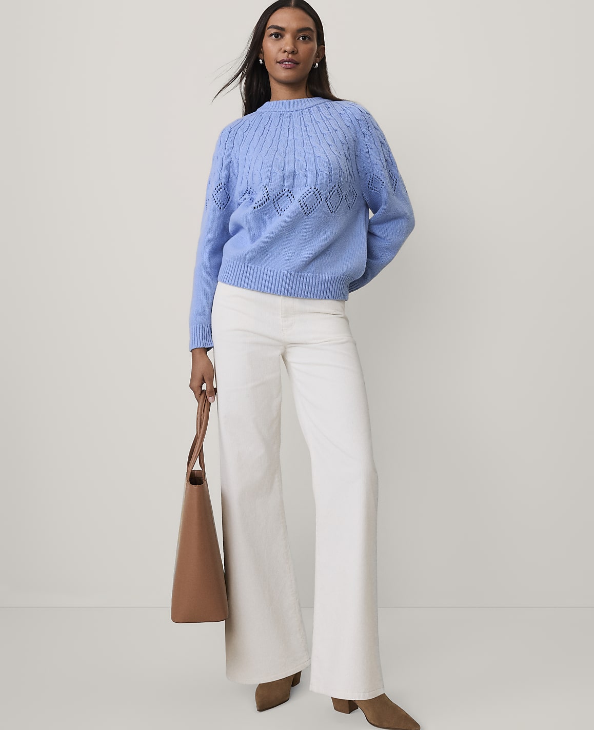 A woman wearing a blue knit sweater, white wide-leg pants, and brown shoes stands against a plain light gray background.