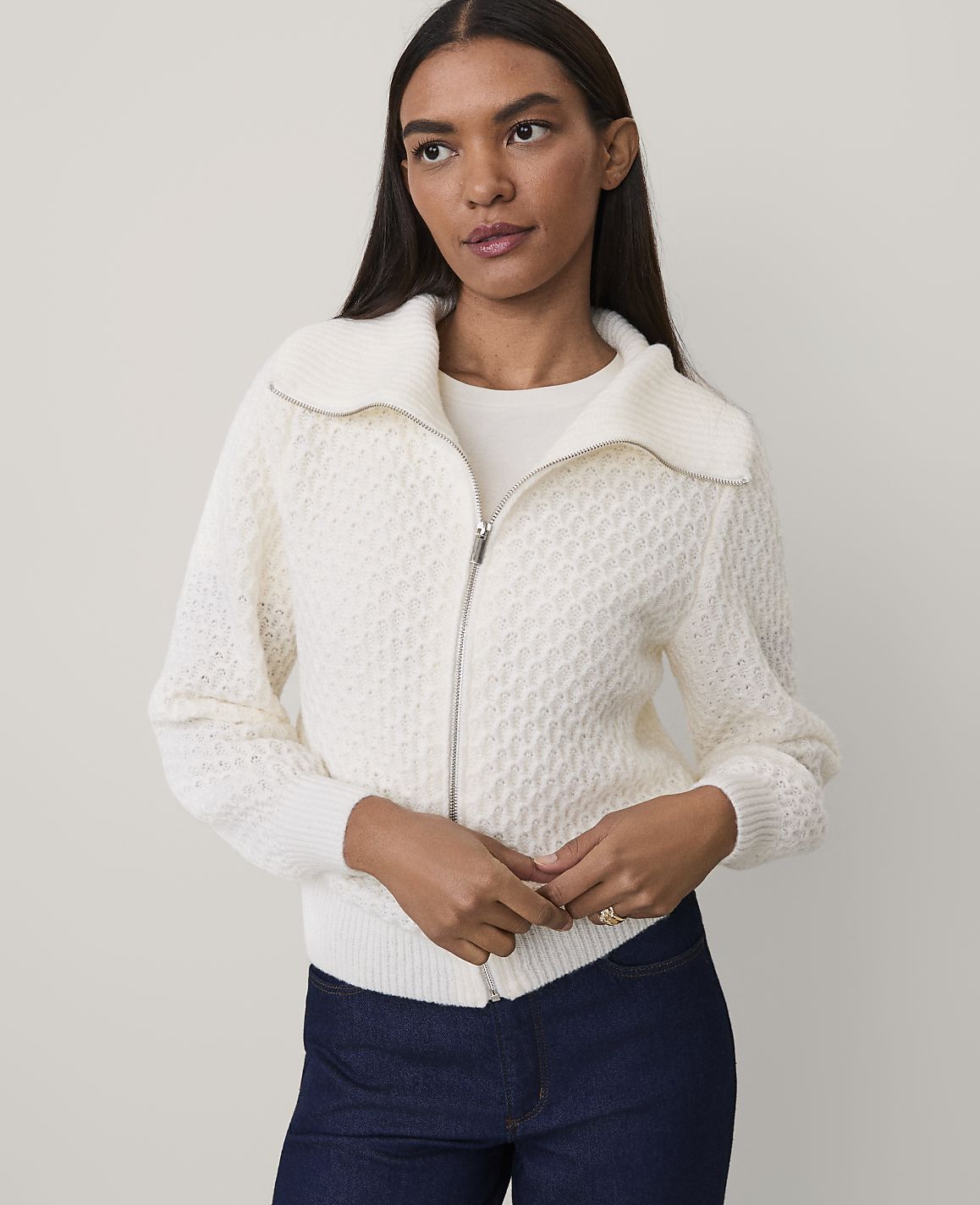 A woman wearing a white textured zip-up sweater and dark blue jeans stands against a plain, light-colored background.