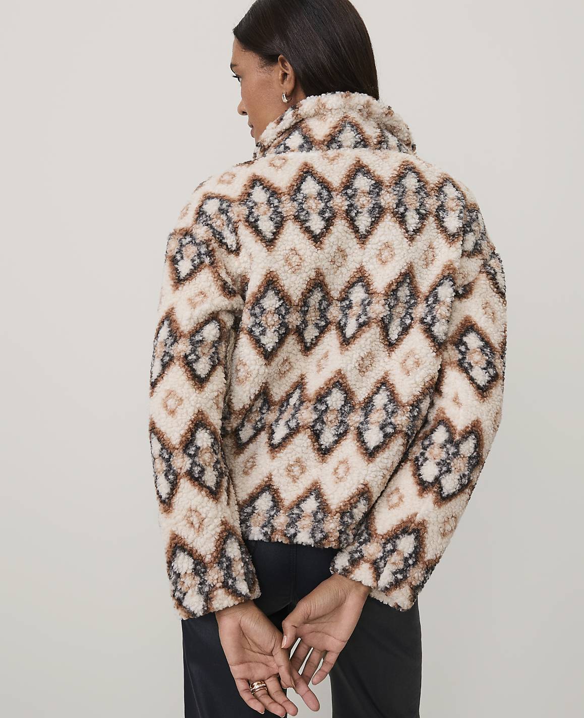 A woman wearing a patterned beige and brown fuzzy jacket stands with her back to the camera against a plain light gray background.