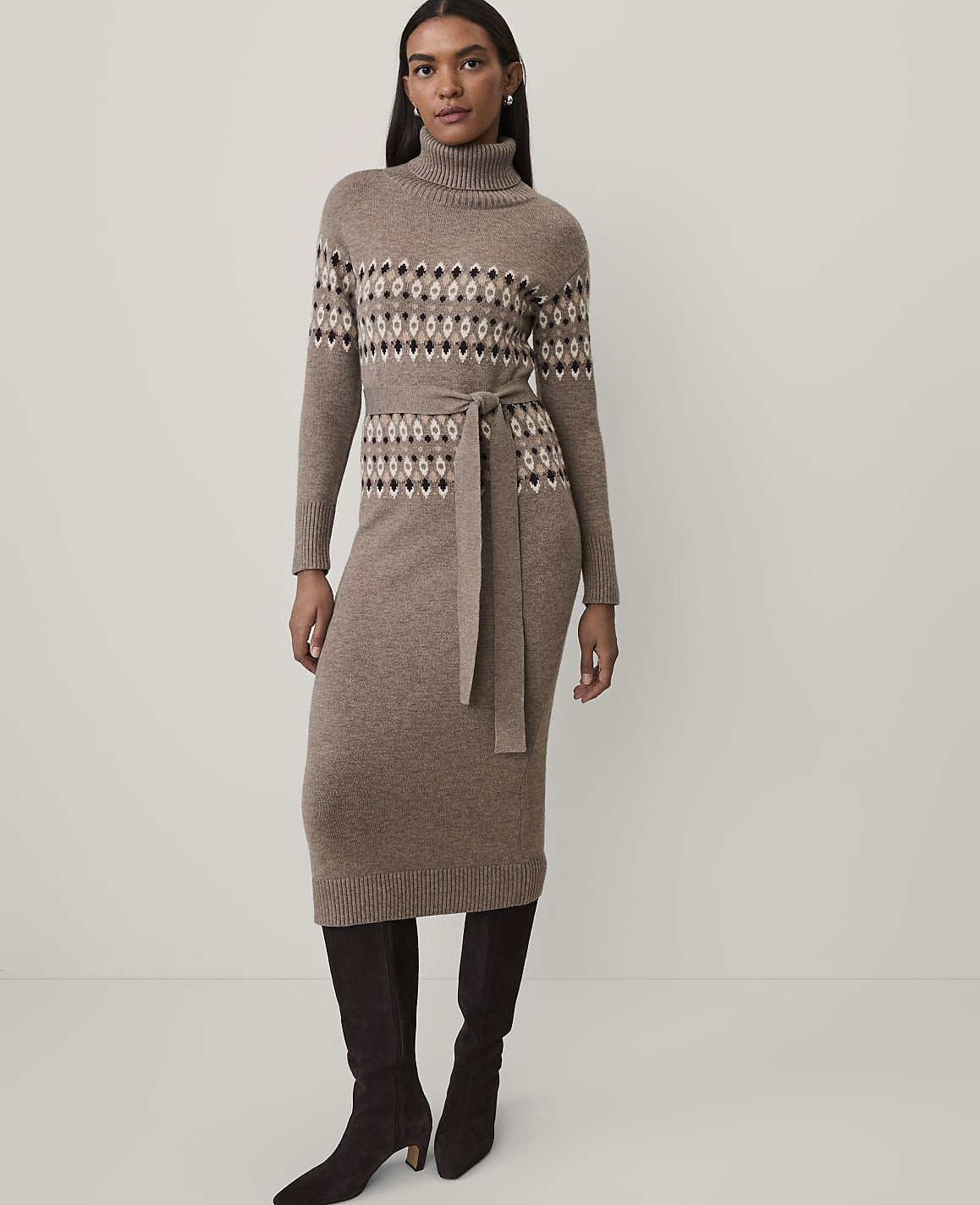 A woman wearing a brown patterned knit dress and dark boots stands against a plain white background.
