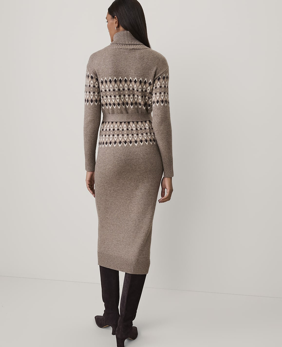 A woman stands with her back to the camera wearing a brown patterned knit dress and dark boots against a plain white background.
