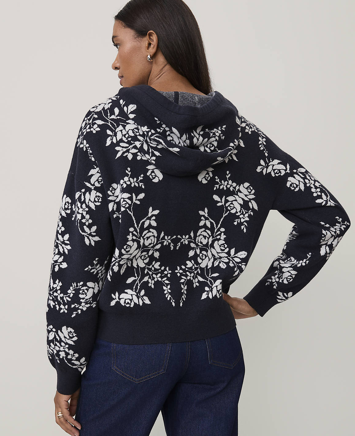 A woman wearing a navy blue floral hooded sweater and dark blue jeans stands against a plain light gray background.
