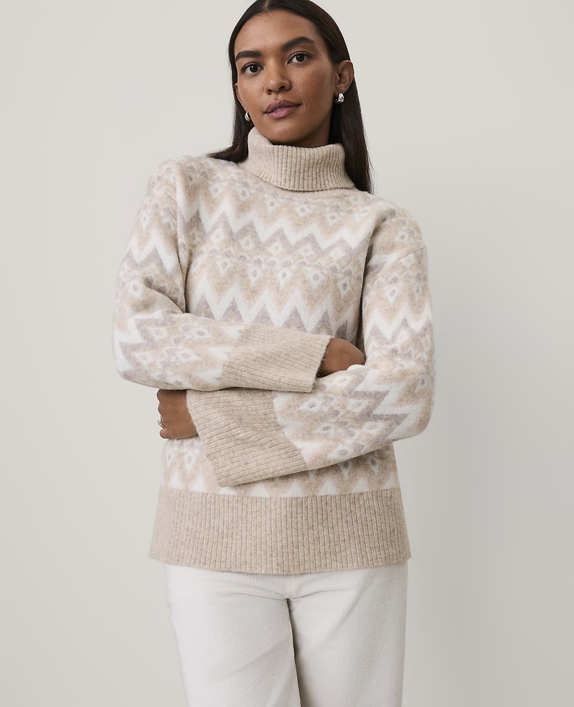 A woman wearing a beige and white patterned turtleneck sweater and white pants stands against a plain light gray background.