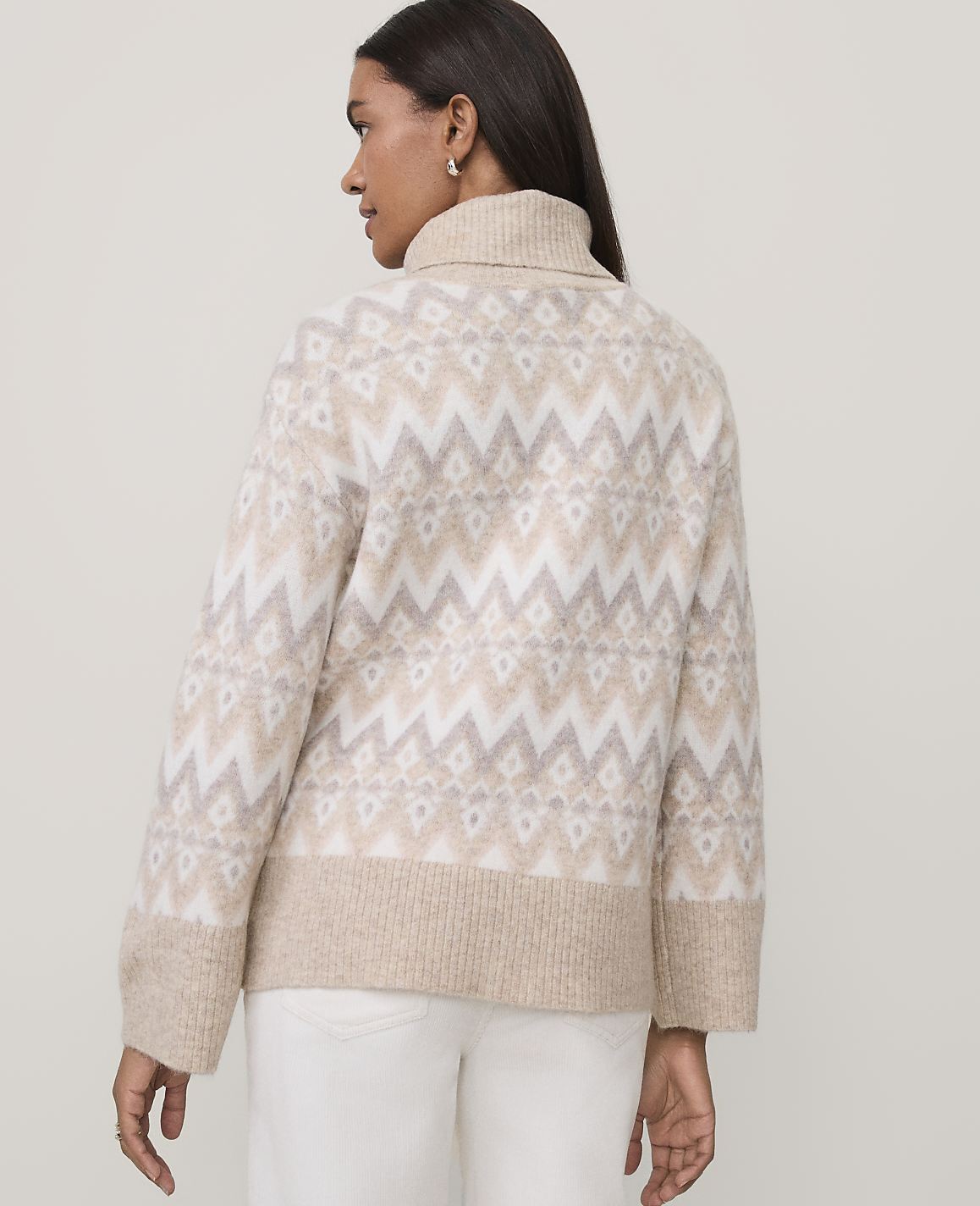 A woman wearing a beige and white patterned turtleneck sweater and white pants stands against a plain light gray background.