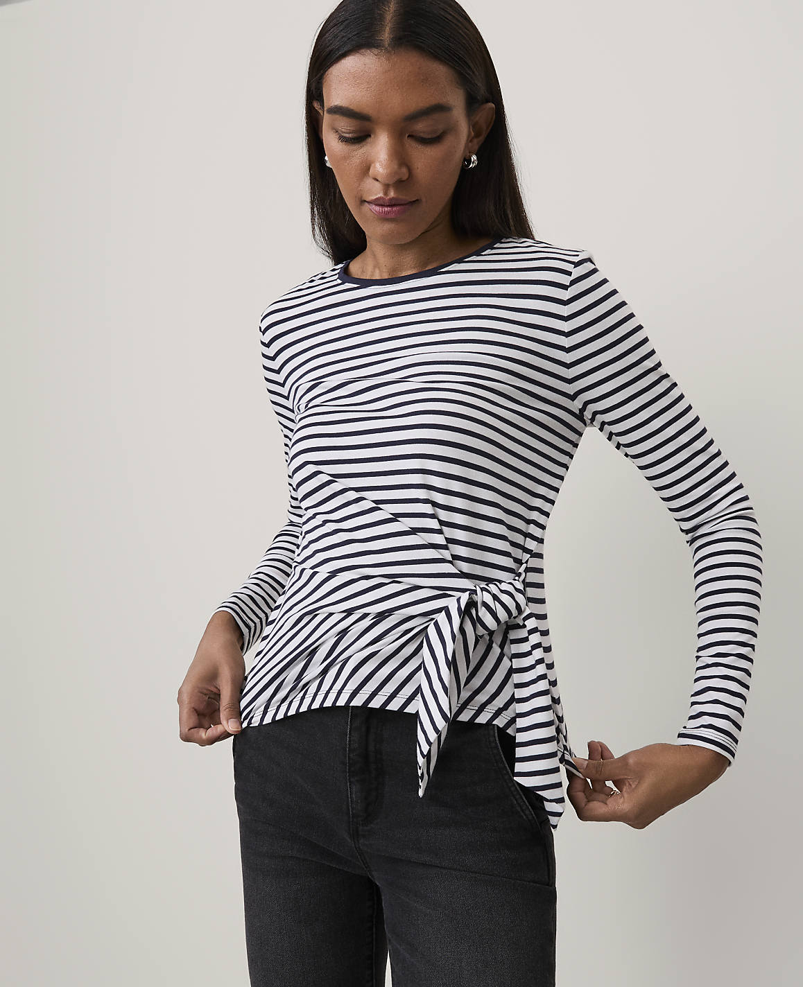 A woman wearing a navy and white striped long-sleeve top with a tie-front detail and black jeans stands against a plain white background.