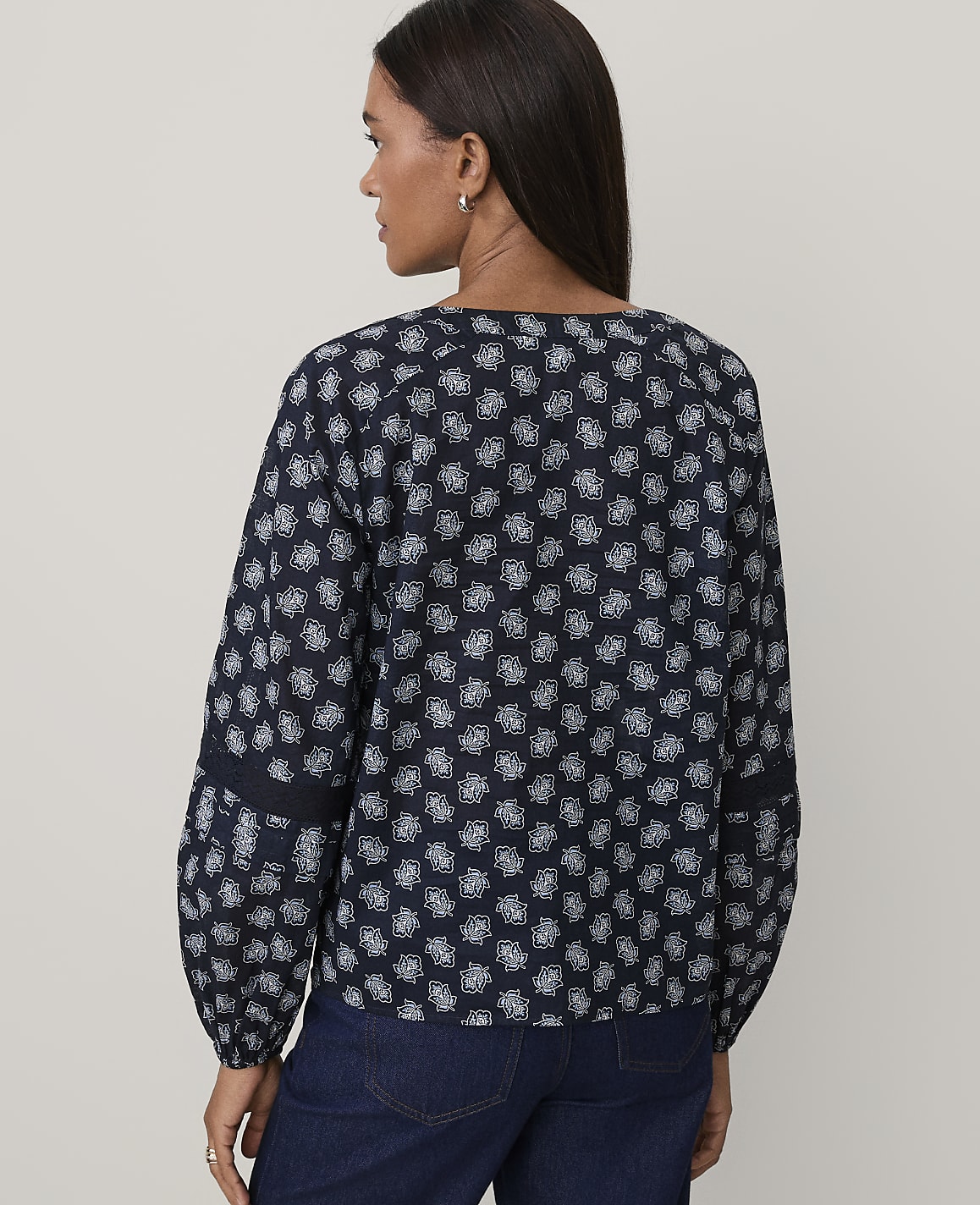 A woman wearing a navy blue patterned blouse and dark blue jeans stands against a plain, light-colored background.