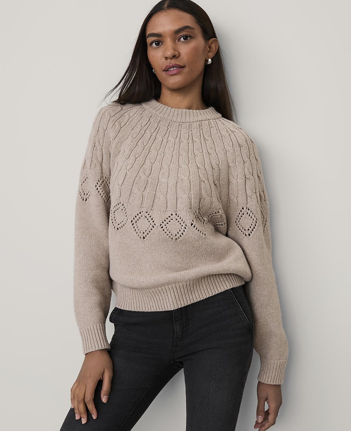 A woman wearing a beige cable-knit sweater and black jeans stands against a plain, light-colored wall.