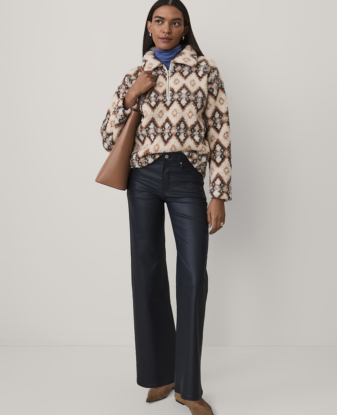 A woman wearing a patterned fuzzy pullover, dark wide-leg trousers, and brown boots stands against a plain light gray background.