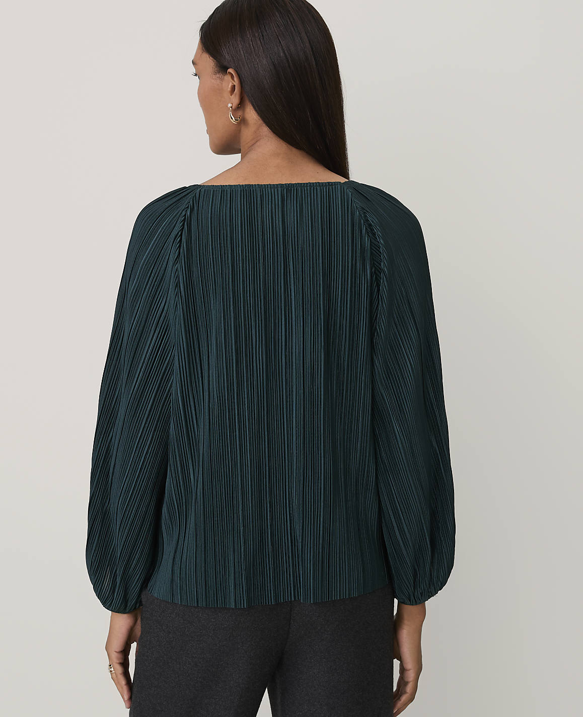 A woman wearing a dark green pleated blouse and charcoal trousers stands against a plain light gray background.