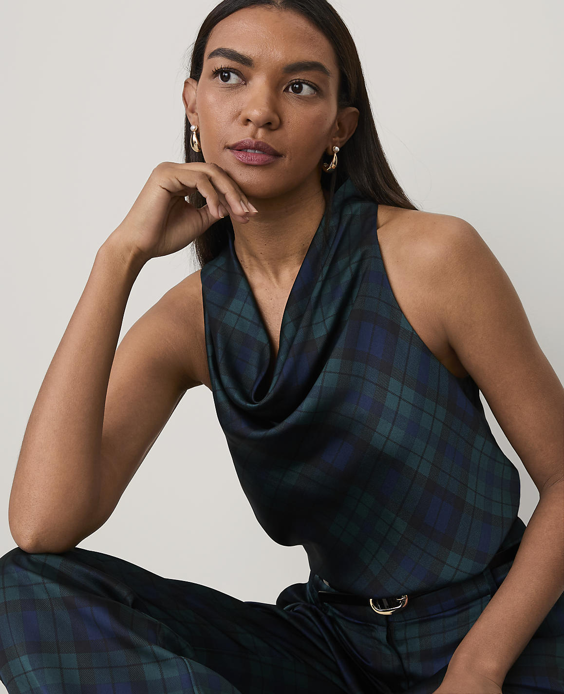 A woman wearing a dark green plaid sleeveless top and matching trousers poses against a plain, light-colored background.