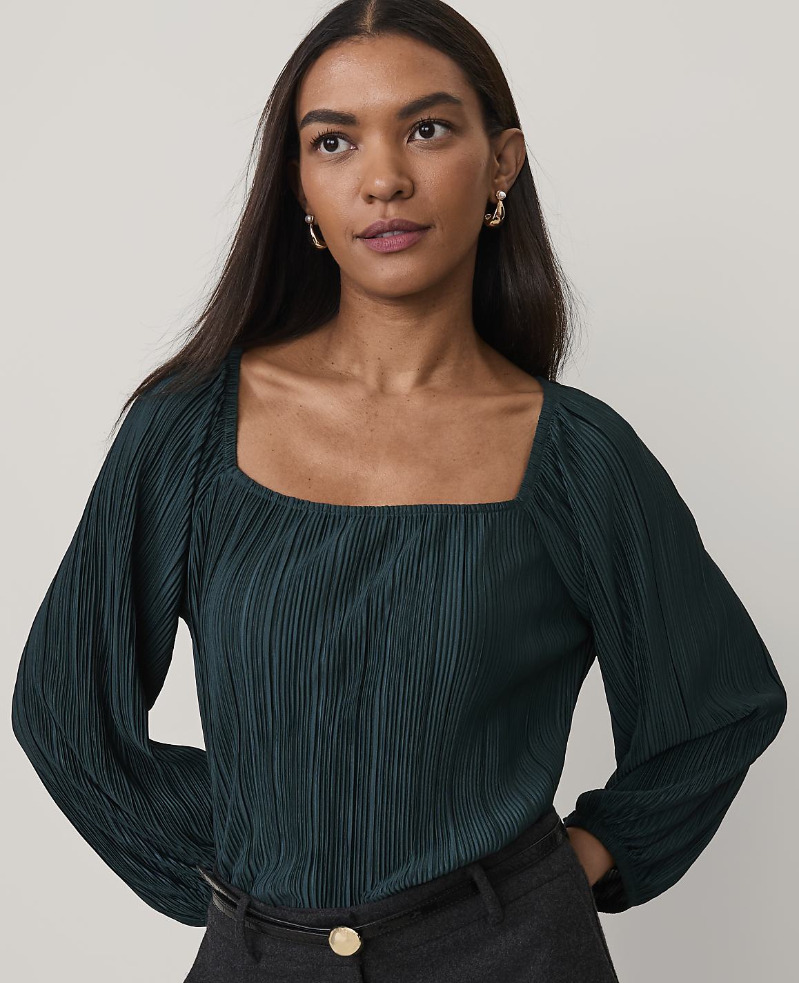 A woman wearing a dark green pleated blouse and grey trousers stands against a plain, light-colored background.
