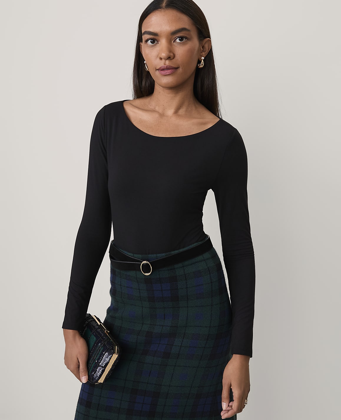 A woman wearing a black long-sleeved top and a green plaid skirt stands against a plain, light-colored background.