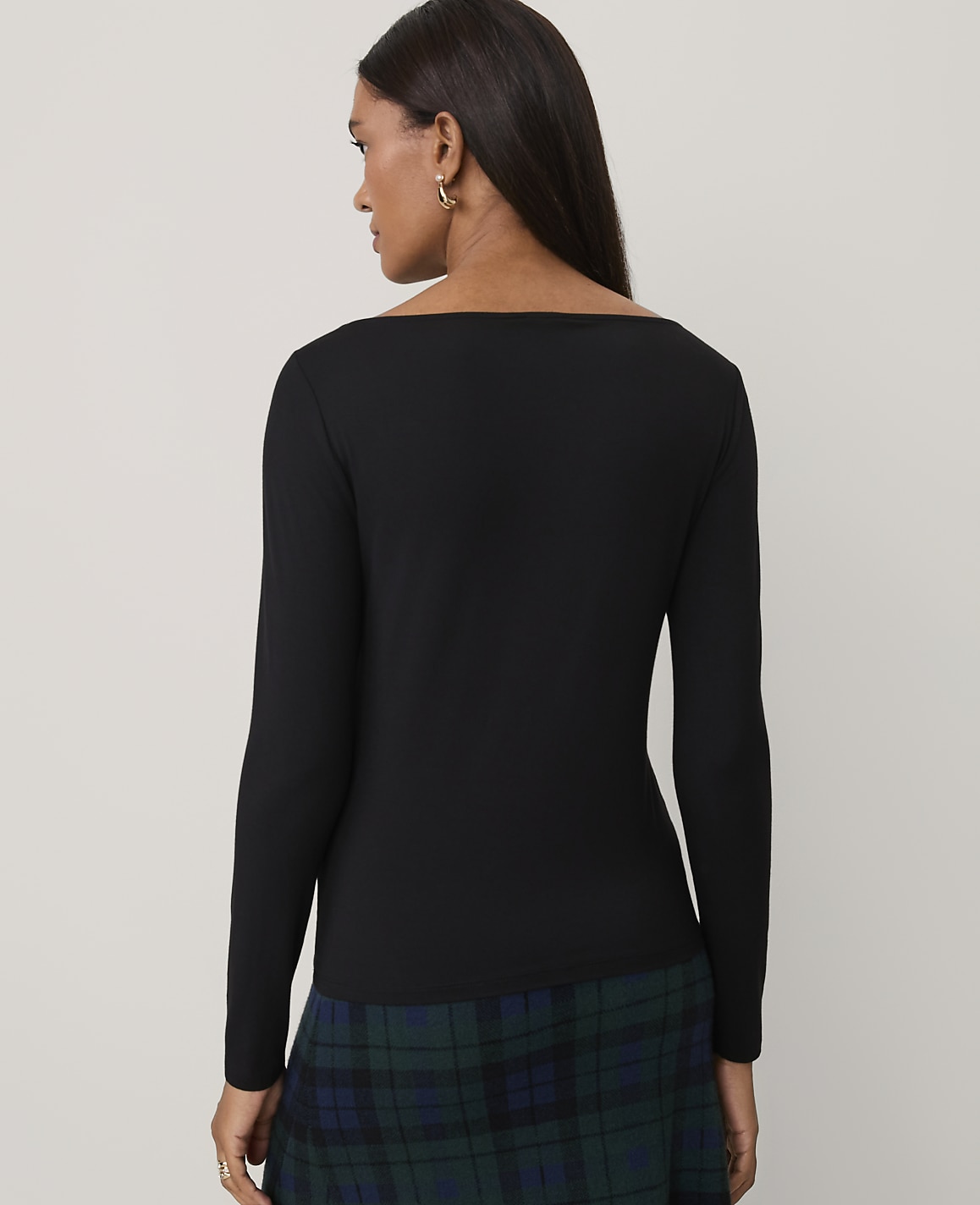 A woman wearing a black long-sleeved top and a green plaid skirt stands against a plain light gray background.
