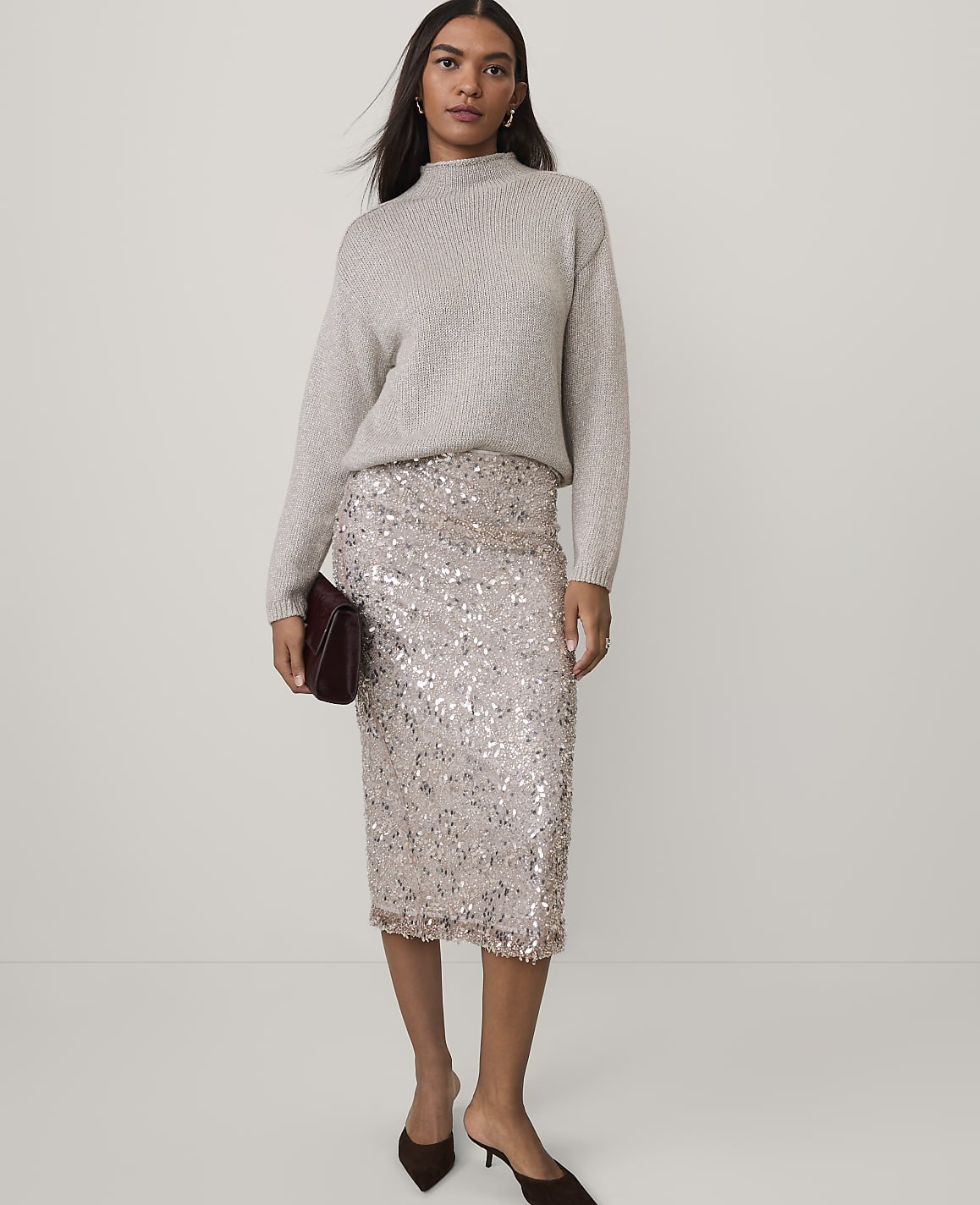A woman wearing a grey sweater and a shimmering sequined skirt stands against a plain, light-colored background.