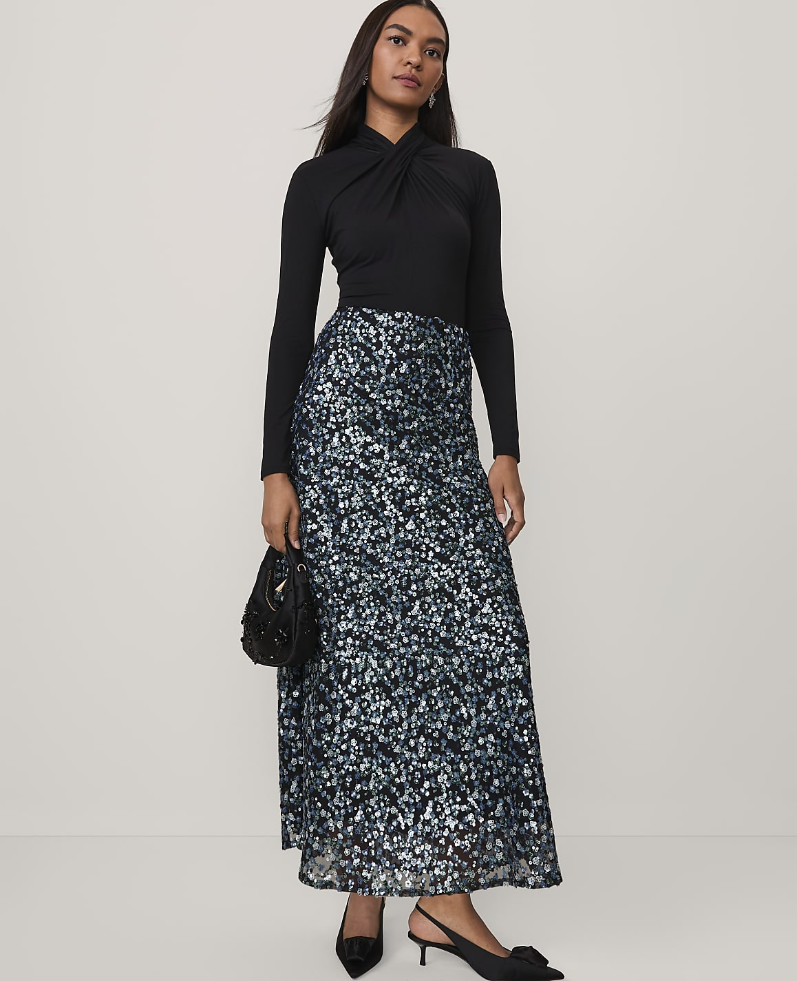 A woman wearing a black long-sleeved top and a shimmering blue and silver sequined skirt stands against a plain, light-colored background.