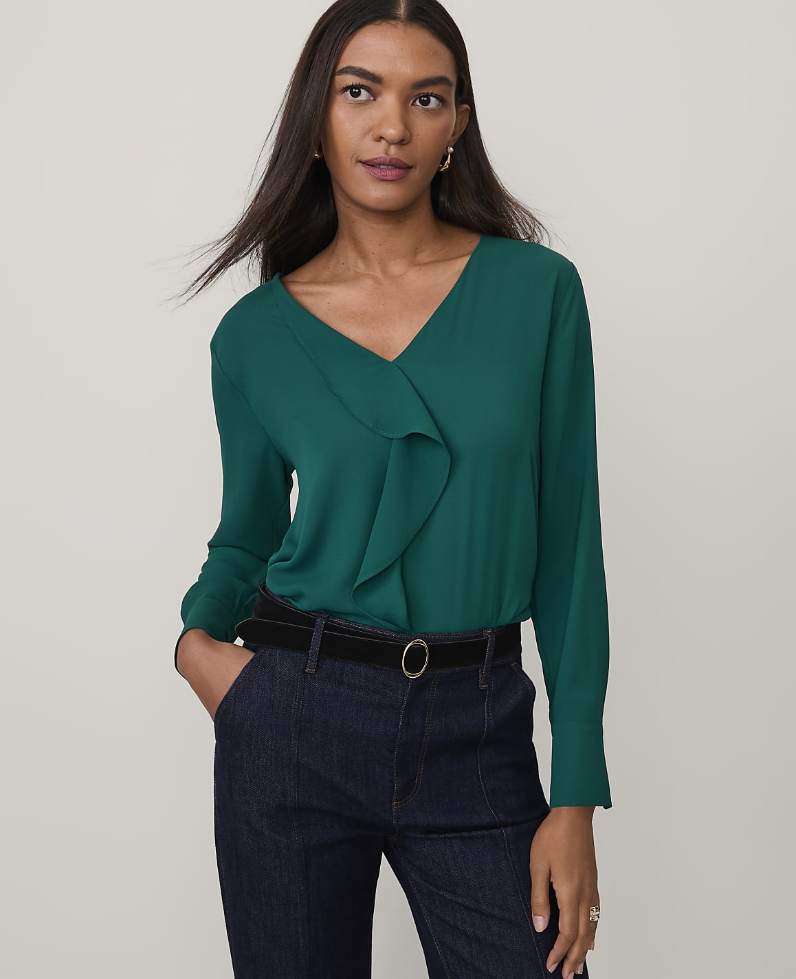 A woman wearing a green ruffle-front blouse and dark blue jeans stands against a plain light gray background.