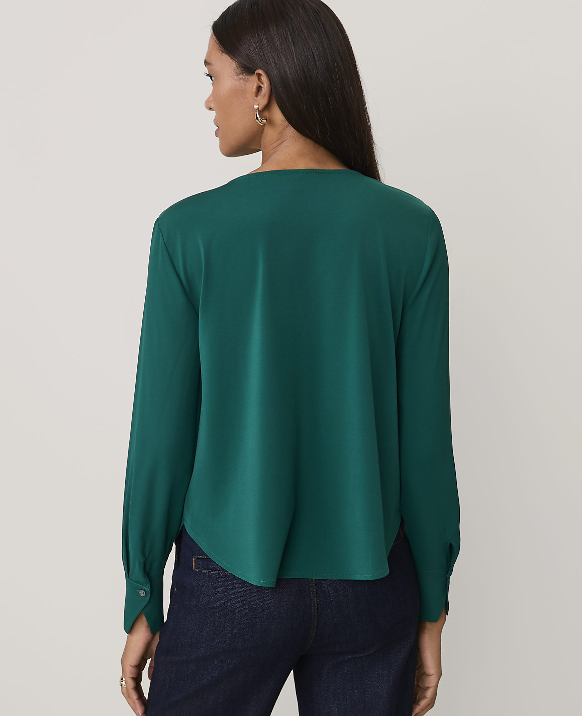 A woman wearing a dark green long-sleeved blouse and navy blue jeans stands against a plain, light-colored background.
