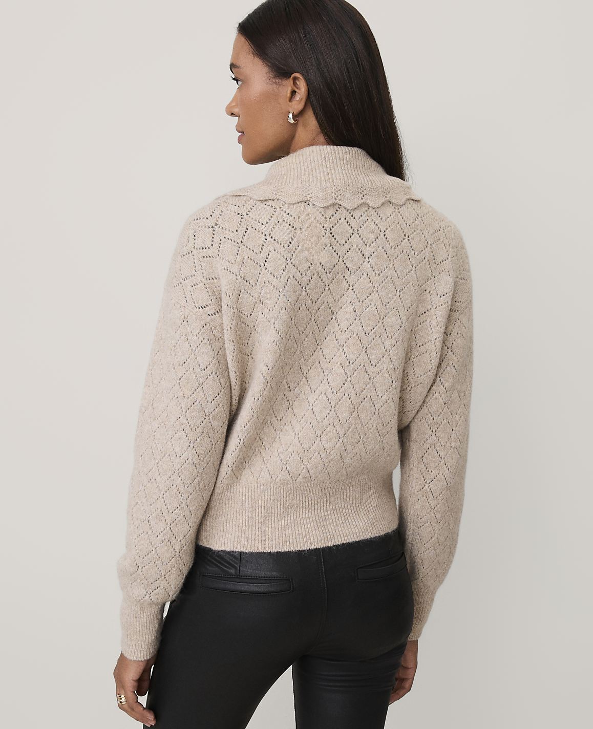 A woman wearing a beige textured sweater and black leather pants stands against a plain light gray background.