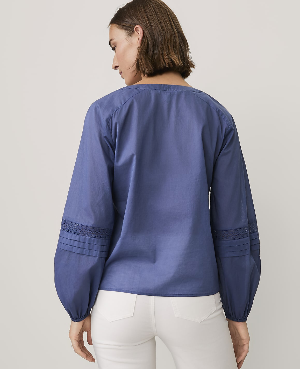 A woman wearing a blue long-sleeved blouse and white pants stands with her back to the camera against a plain light gray background.