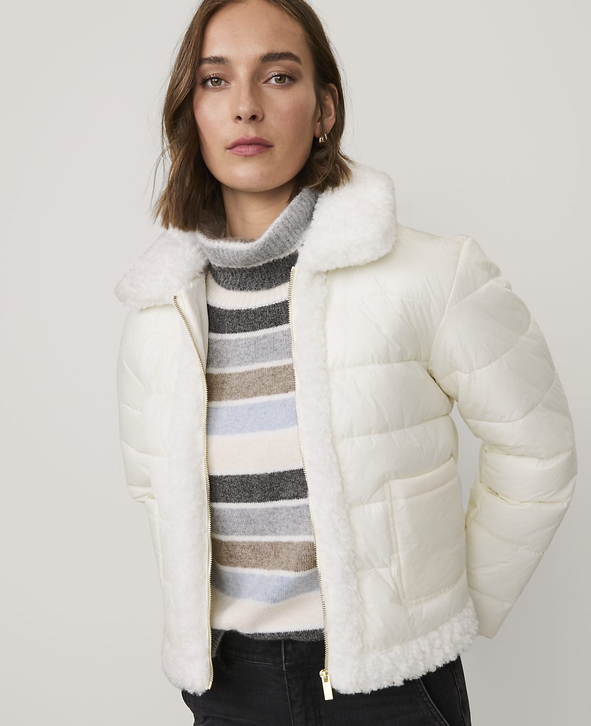 A woman wearing a white puffer jacket and a striped sweater stands against a plain, light-colored background.