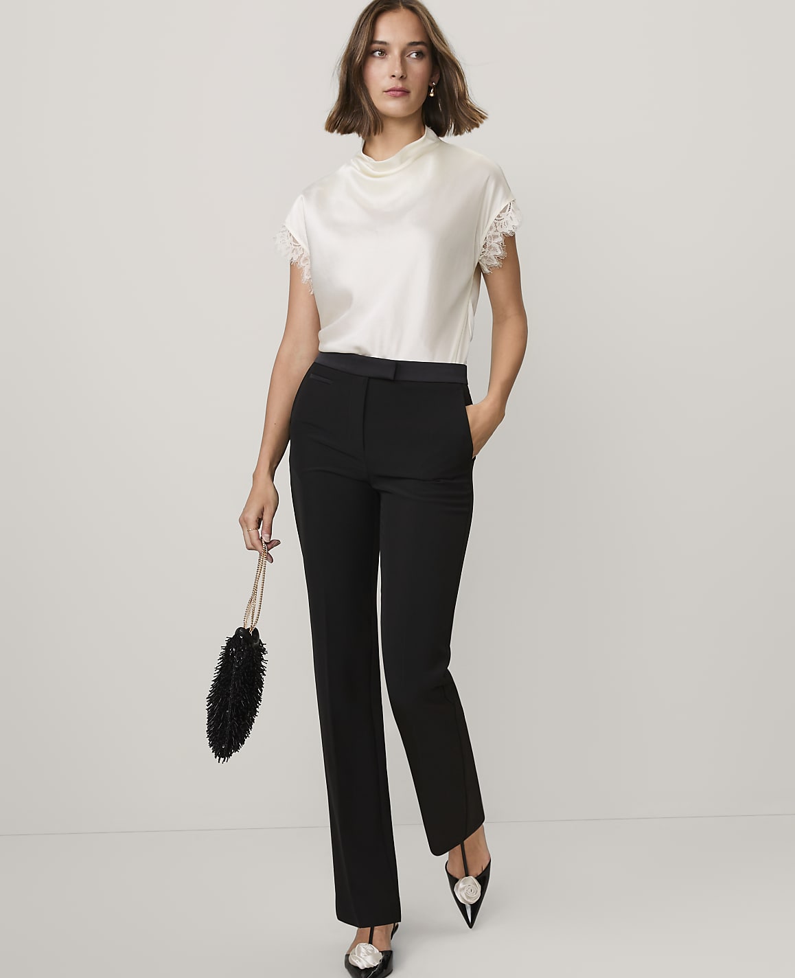 A woman wearing a white lace-trimmed top and black trousers stands against a plain white background.