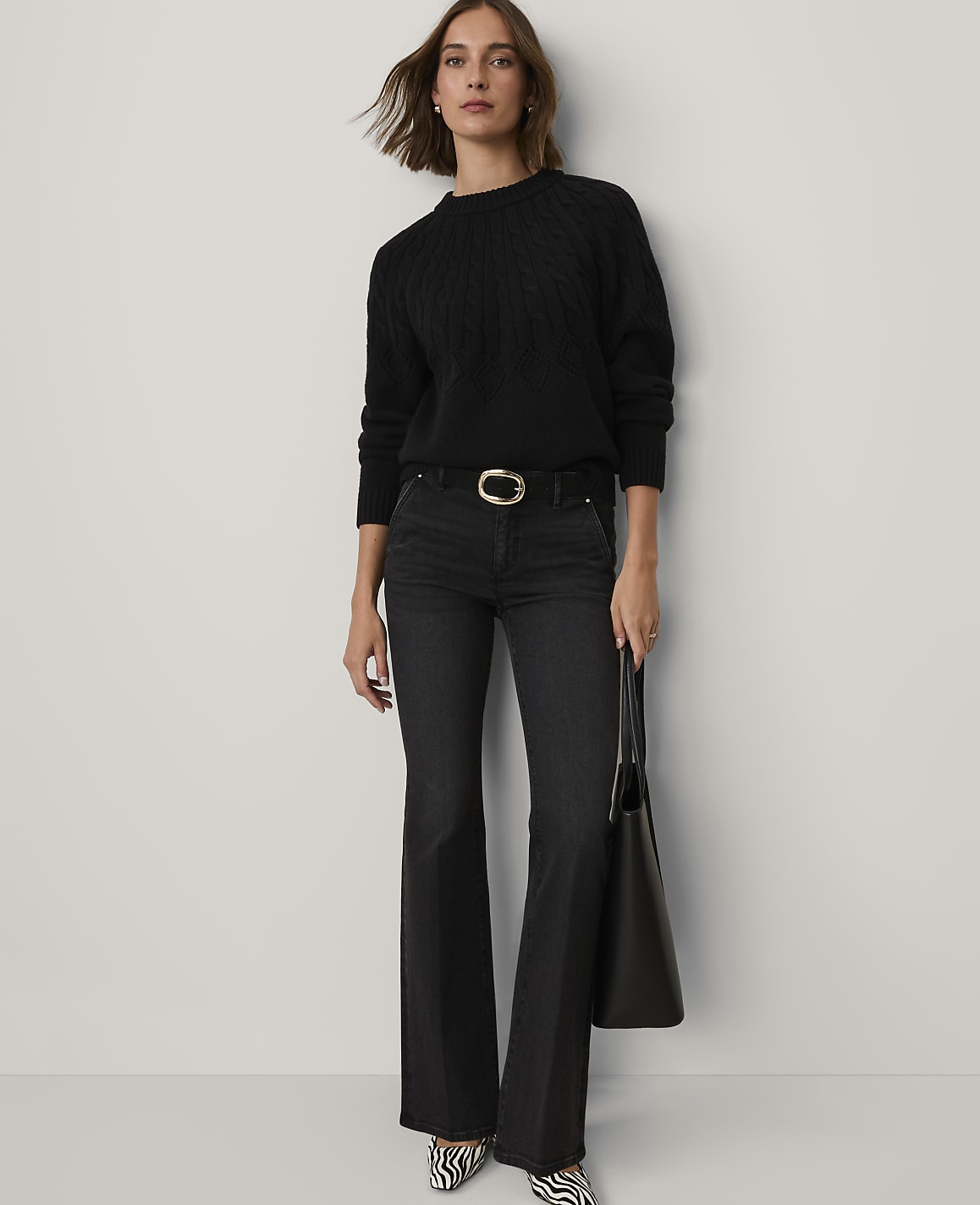 A woman wearing a black cable-knit sweater, dark flared jeans, and zebra-print shoes stands against a plain, light-colored background.