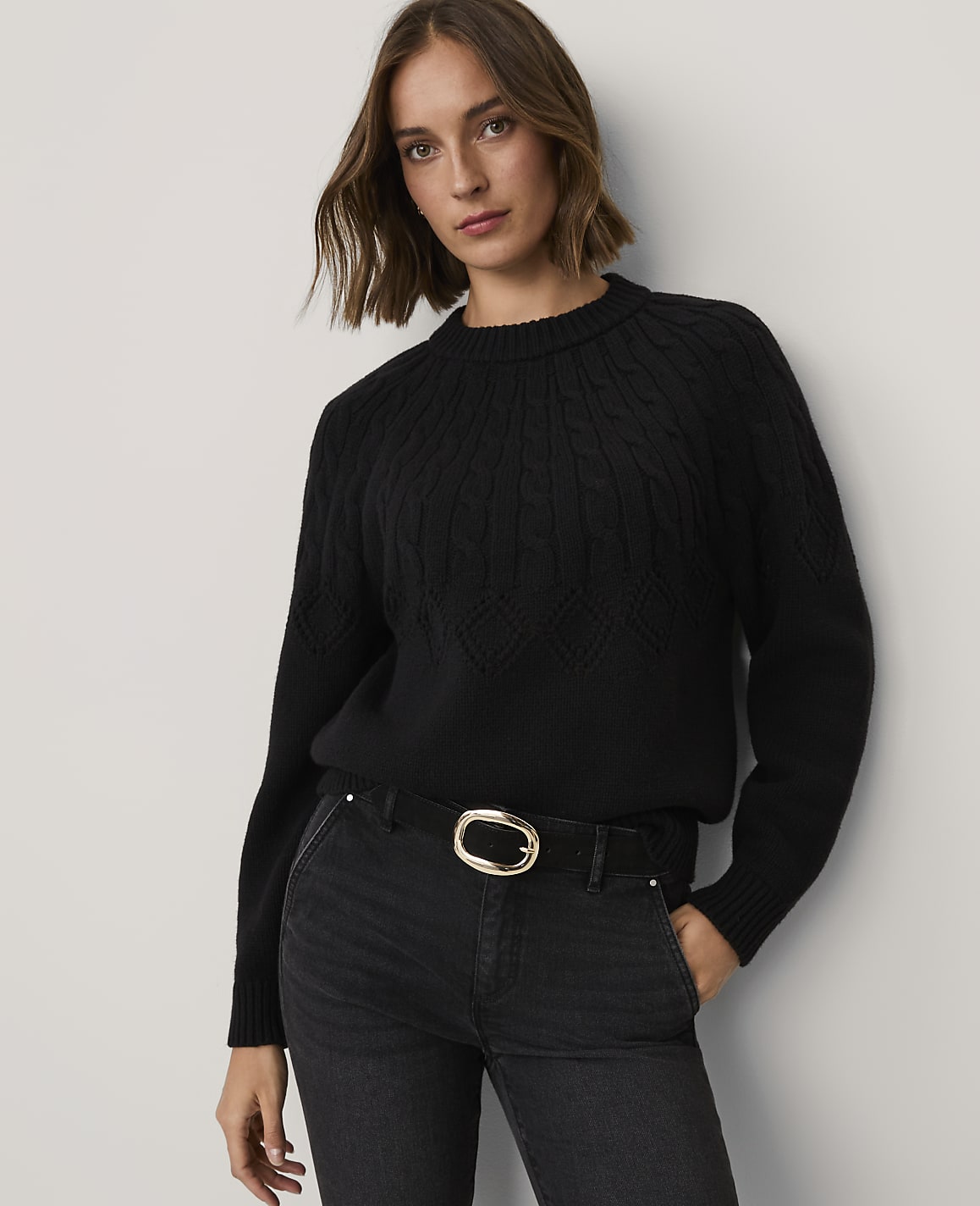 A woman wearing a black cable-knit sweater and black jeans with a gold buckle belt stands against a plain white wall.