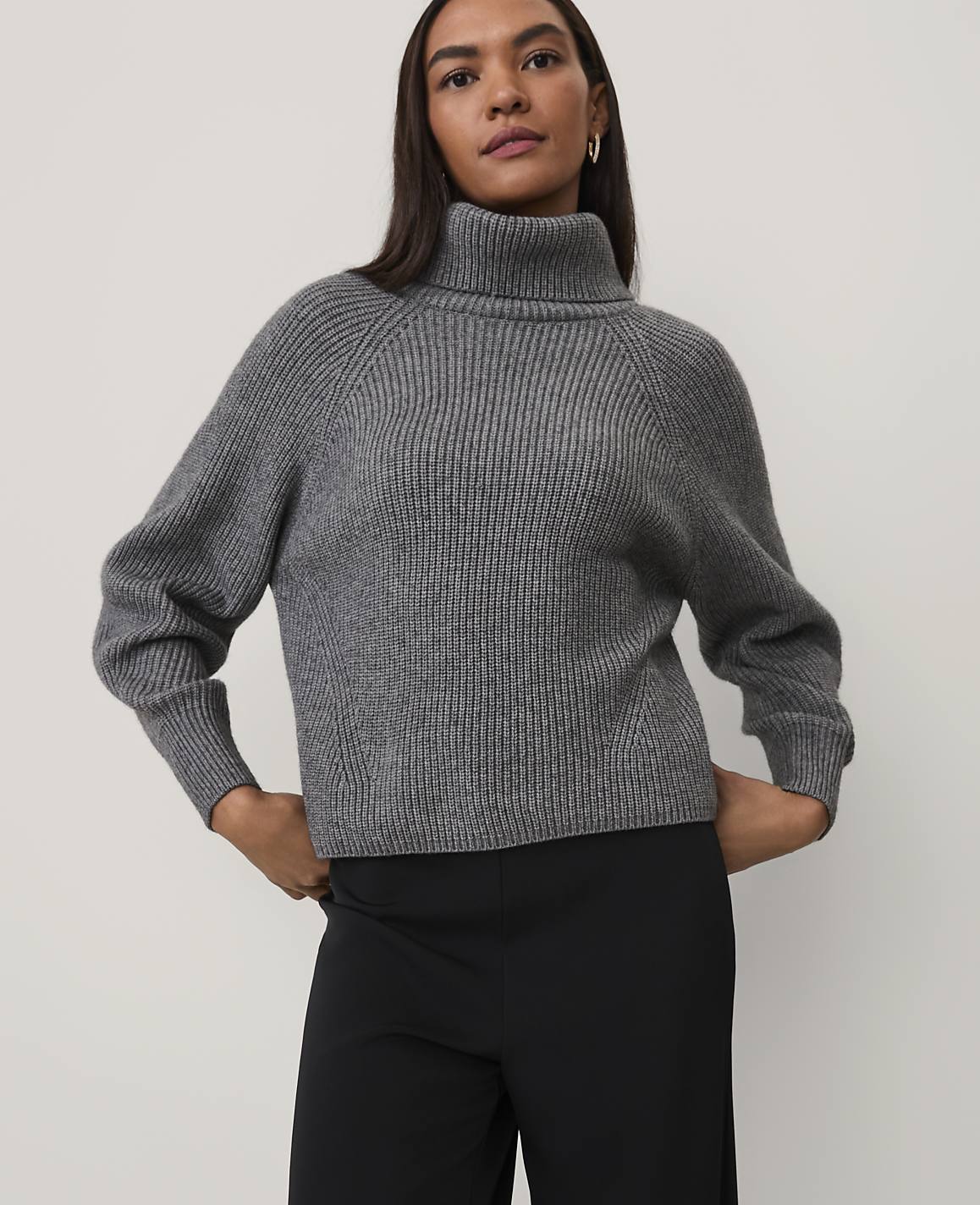 A woman wearing a grey ribbed turtleneck sweater and black trousers stands against a plain light grey background.