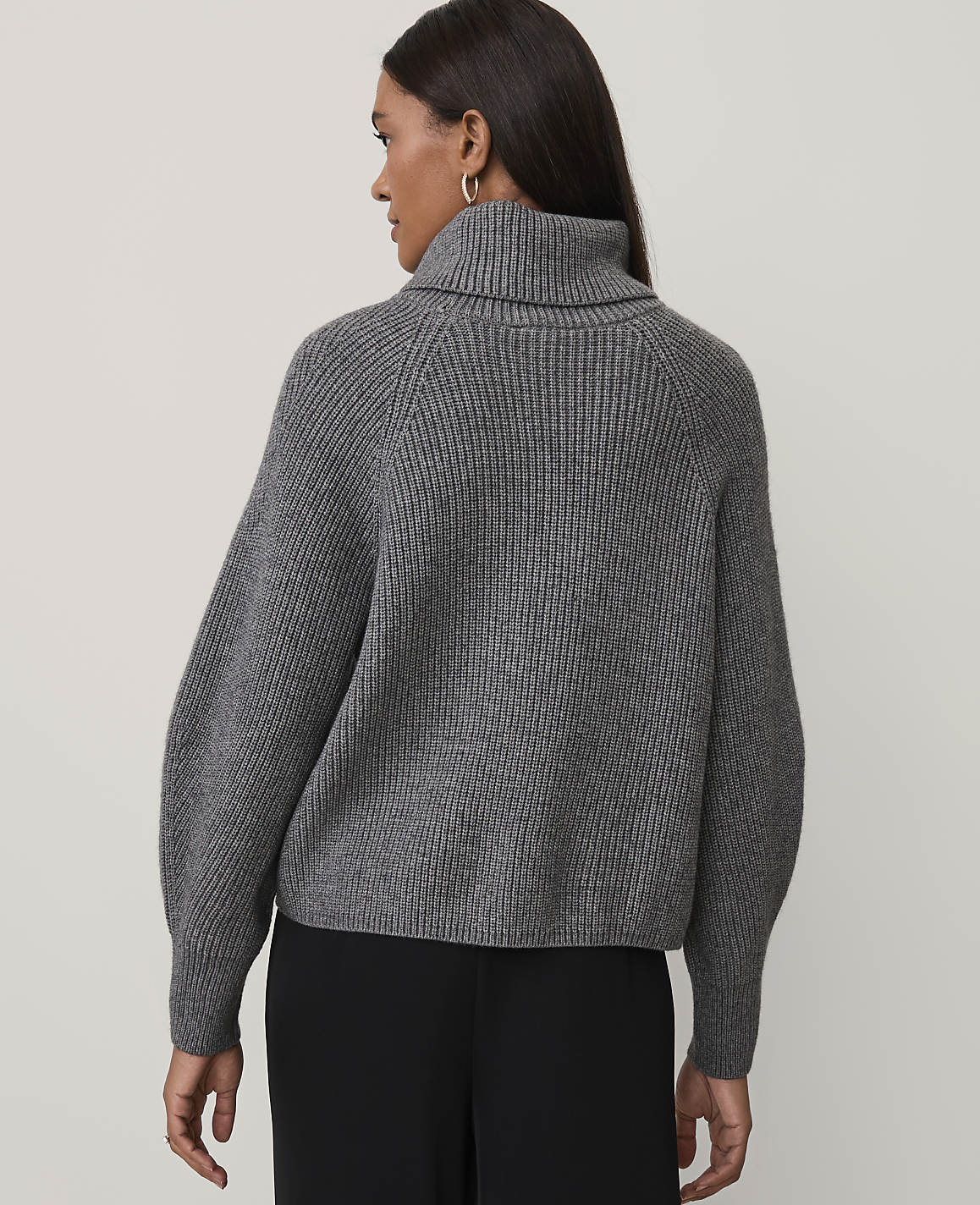 A woman wearing a grey ribbed turtleneck sweater and black trousers stands against a plain light-colored background.