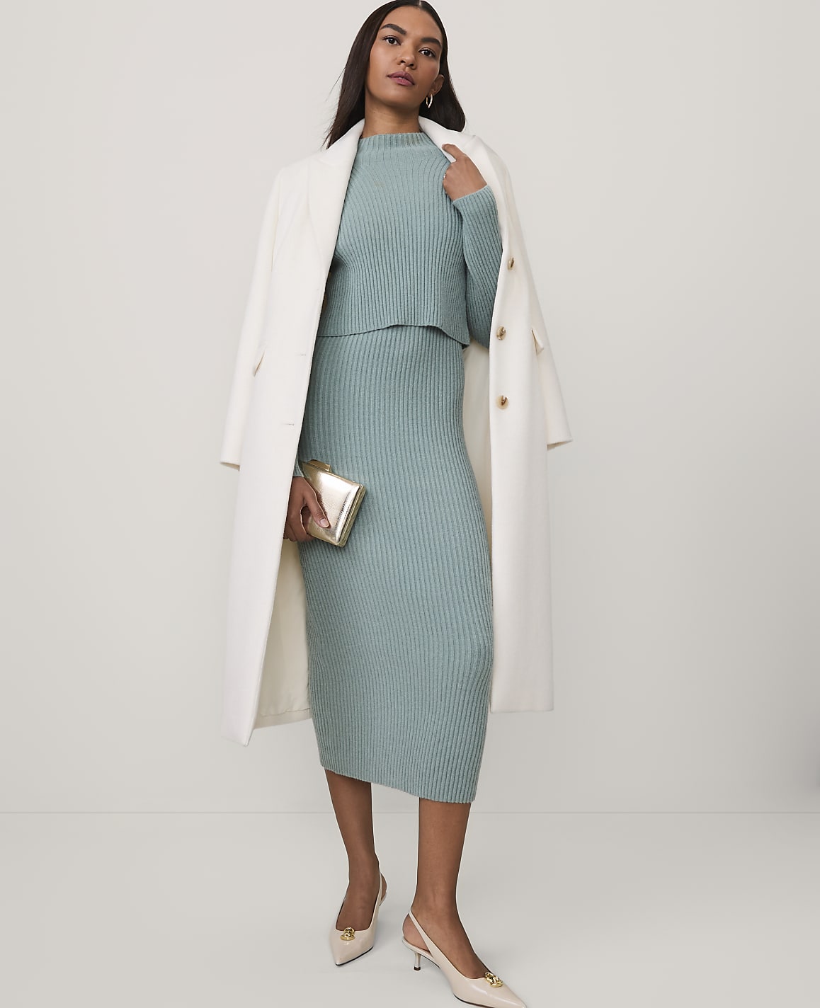 A woman wearing a sage green ribbed knit set and a white coat stands against a plain white background.
