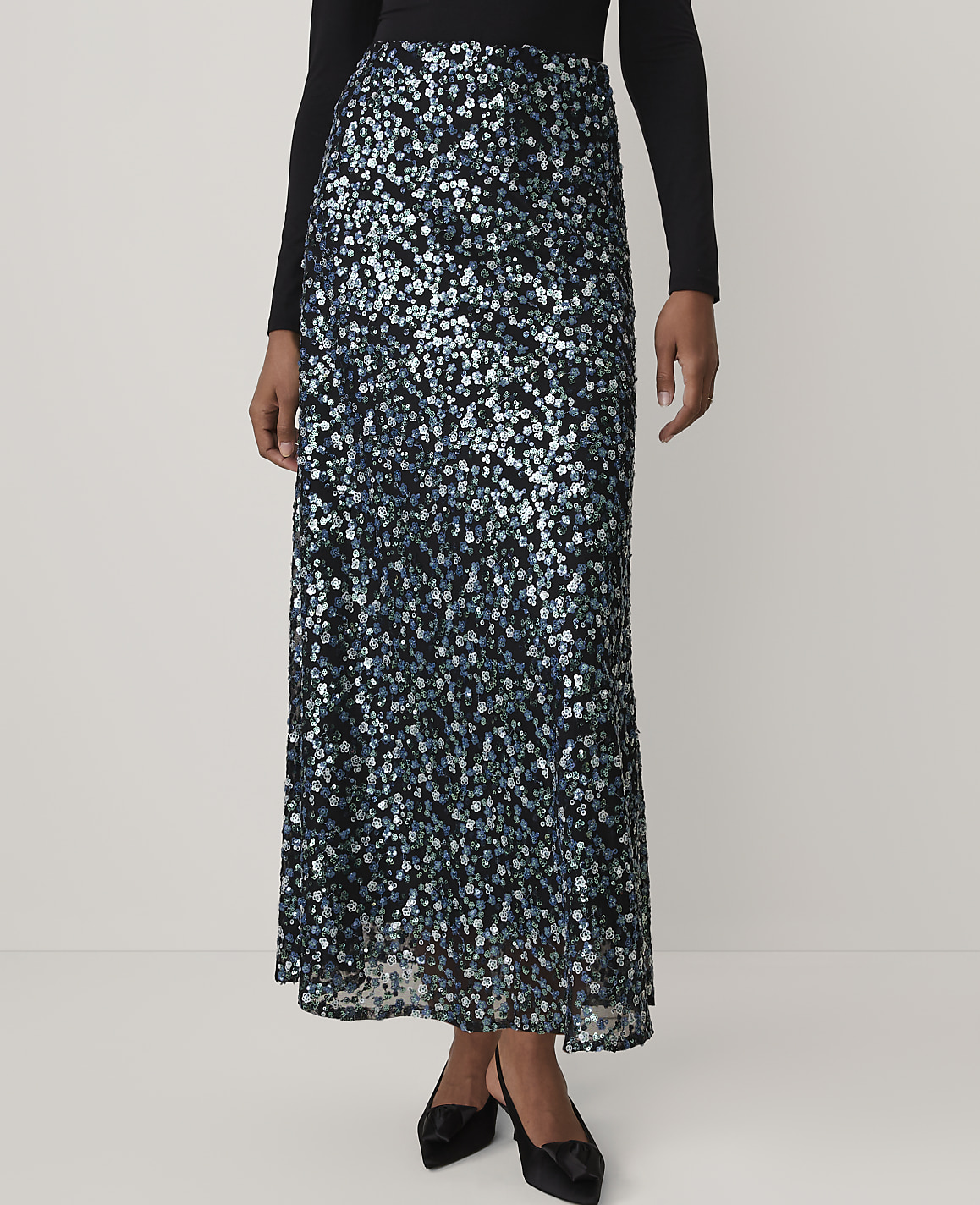A person wears a long, shimmering blue and silver sequined skirt and a black top against a plain, light-colored background.