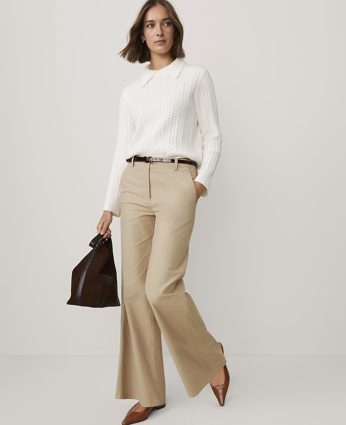 A woman wearing a white cable-knit sweater, tan flared trousers, and brown shoes stands against a plain light gray background.
