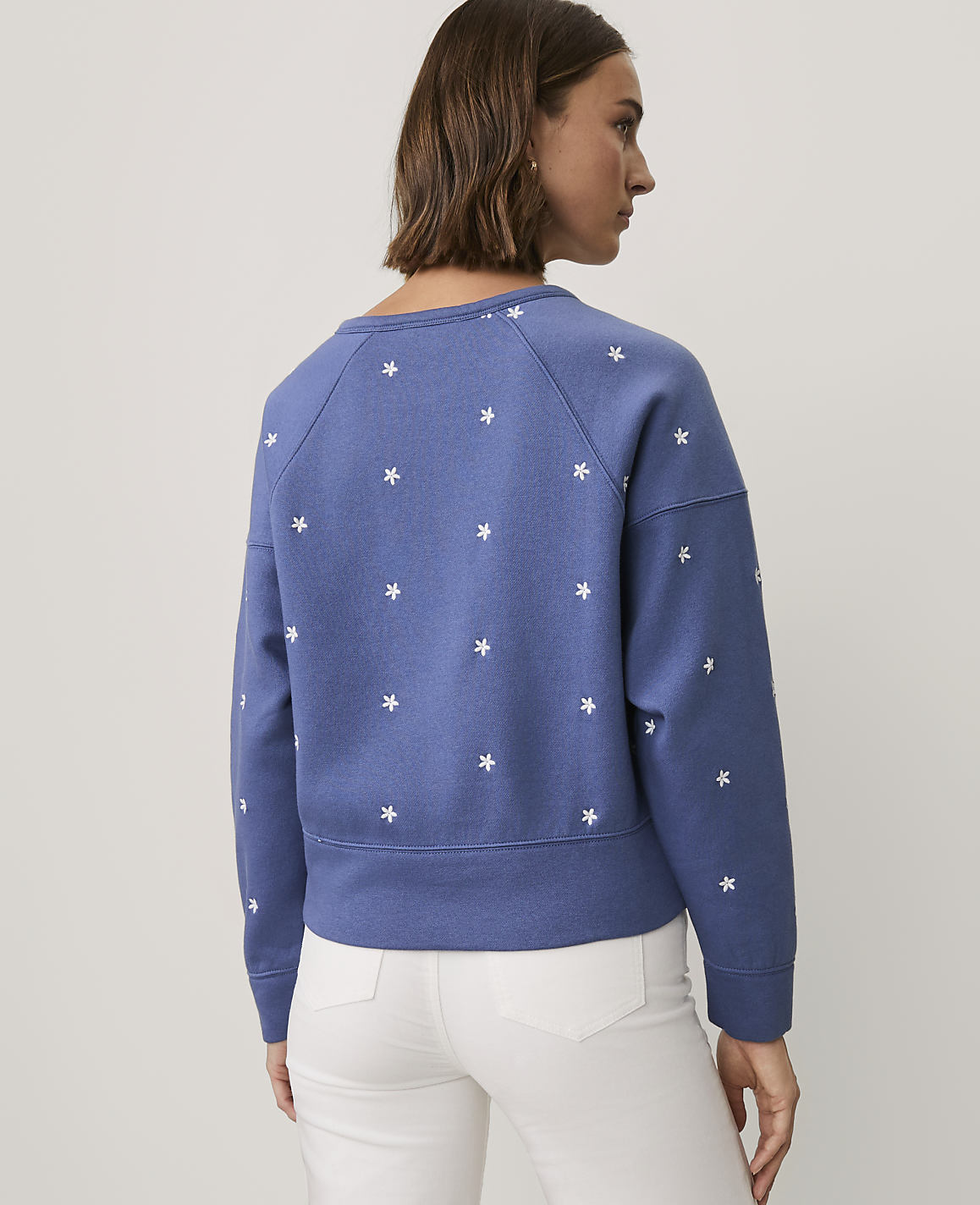 A woman wearing a blue floral-patterned sweatshirt and white pants stands against a plain light gray background.