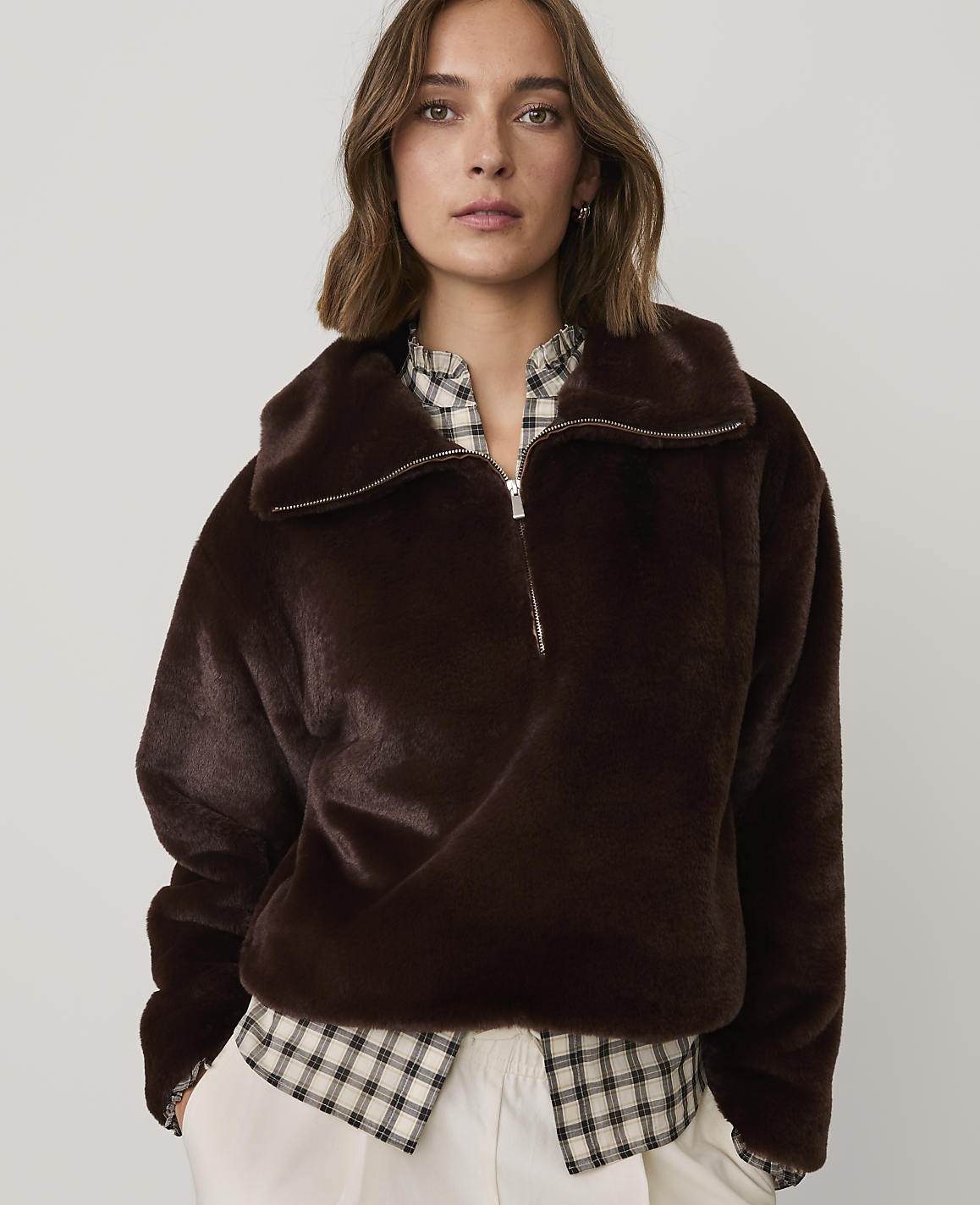 A woman wearing a dark brown faux-fur jacket over a checkered shirt and white trousers stands against a plain light gray background.