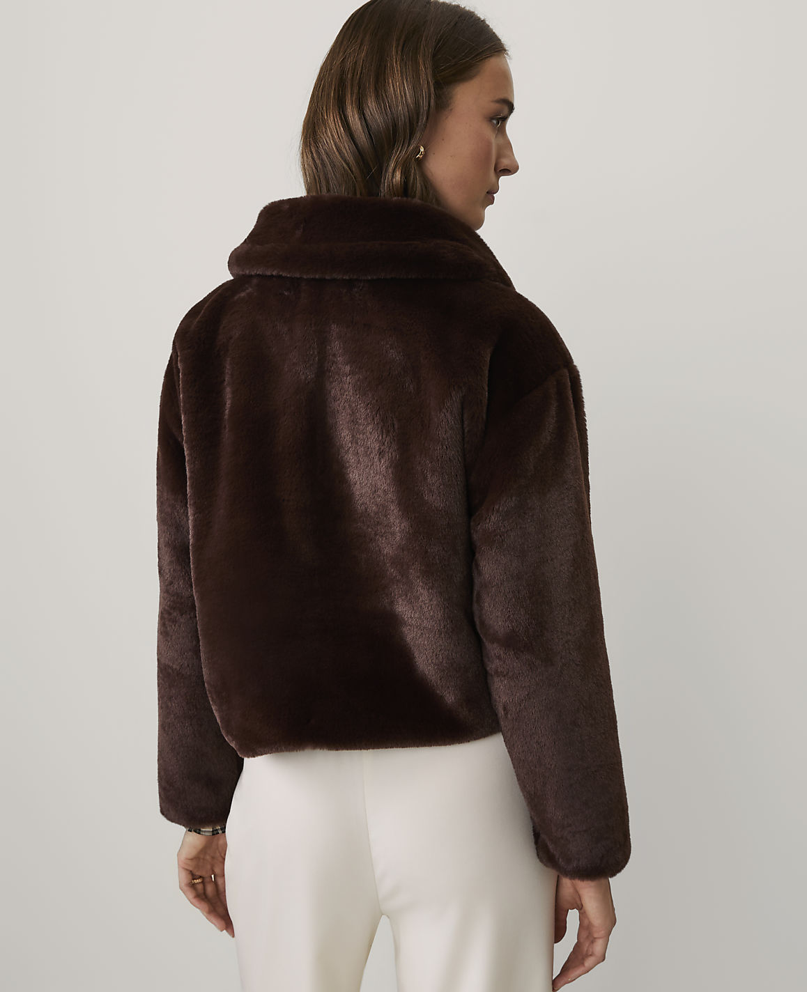 A woman wearing a dark brown faux fur jacket and white trousers stands against a plain white background.