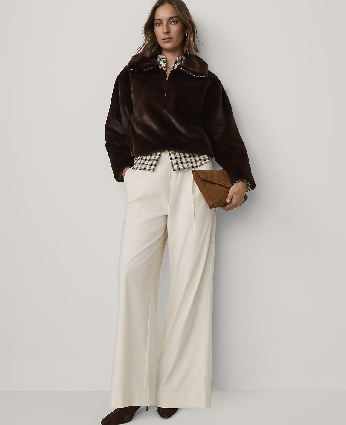 A woman wearing a brown faux-fur jacket, a checkered shirt, and wide-leg white trousers stands against a plain white background.