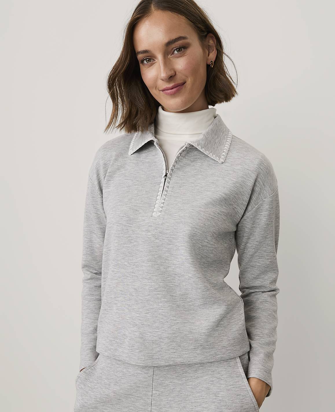 A woman wearing a grey quarter-zip pullover and matching pants stands against a plain, light-colored background.