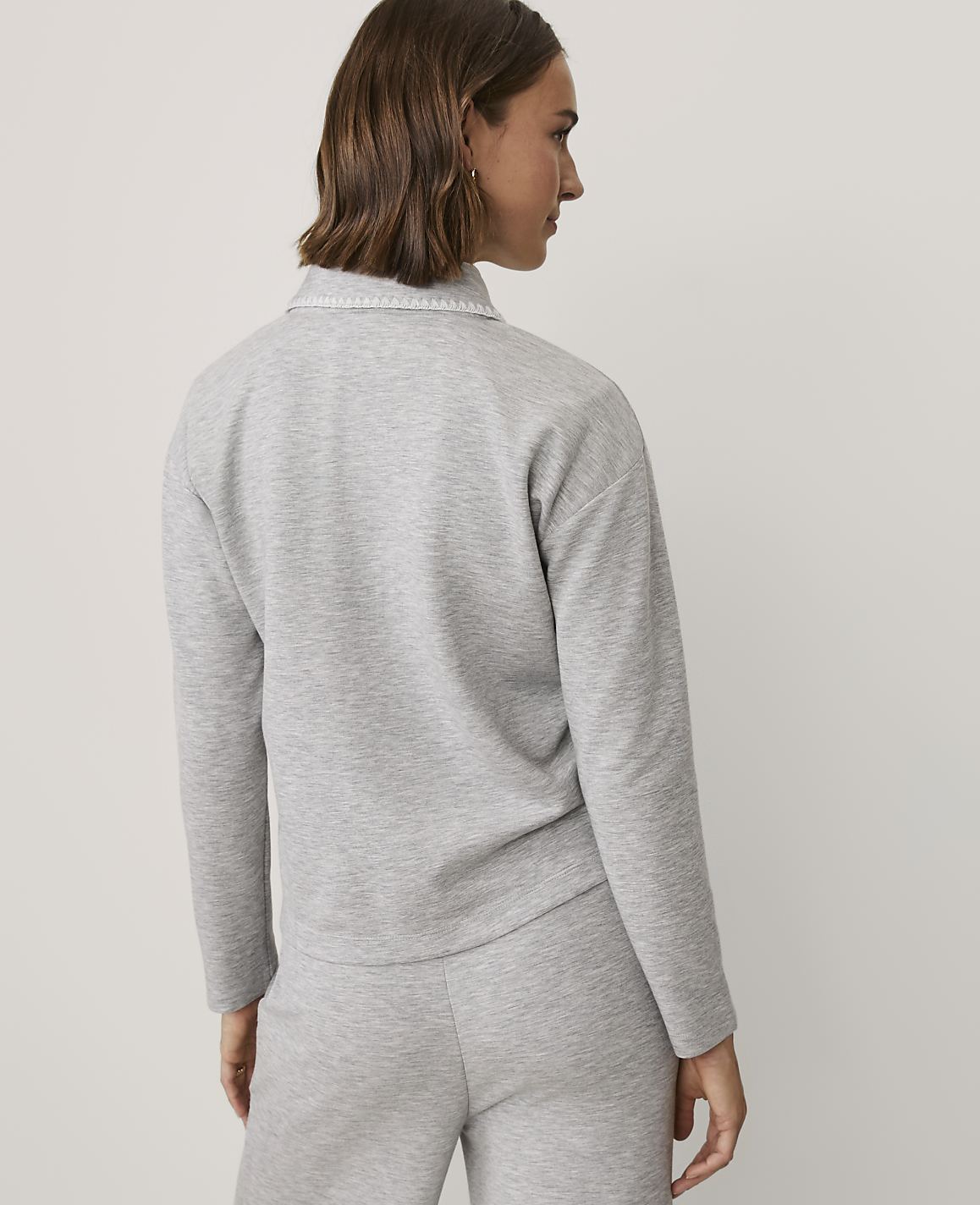 A woman with brown hair wearing a grey matching sweatshirt and pants set stands against a plain white background.