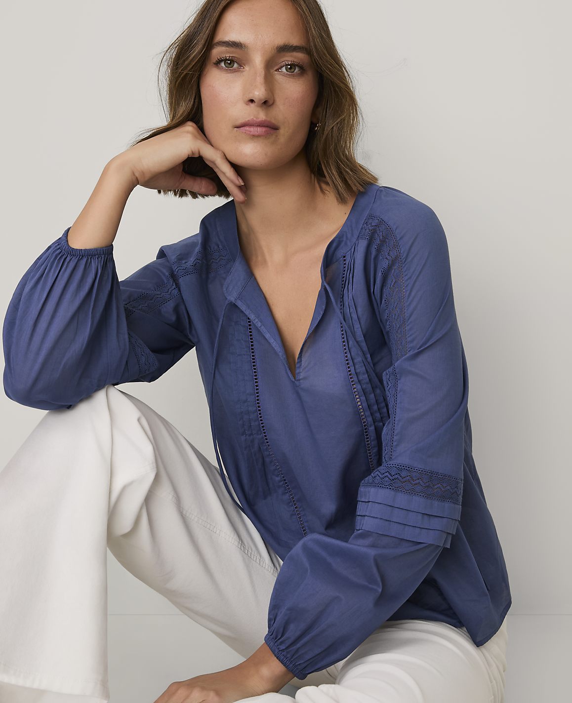 A woman wearing a blue long-sleeved blouse and white pants poses against a plain white background.